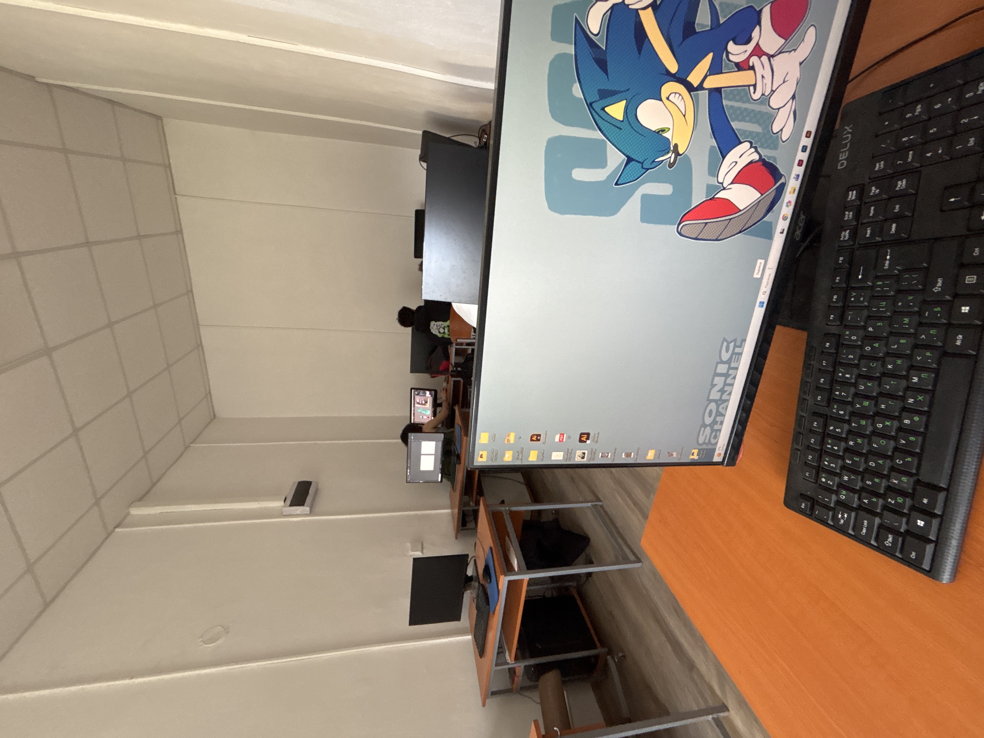 This image, rotated clockwise, depicts an indoor computer lab or classroom in Troyan, Bulgaria. In the foreground, a computer monitor displays a vibrant Sonic the Hedgehog wallpaper, with the character prominently featured and the text "SONIC CHANNEL" visible. The monitor has an "Acer" brand logo on its lower bezel. To the right, a black "DELUX" branded keyboard with Cyrillic characters is positioned on a wooden desk.
In the midground and background, several individuals are seated at their respective computer workstations, appearing to be working or studying. One person with dark hair and a dark top is clearly visible facing their monitor. The workstations consist of wooden-topped desks with metal frames and chairs. The room has light-colored walls, a ceiling with acoustic tiles, and artificial lighting fixtures. A small, white rectangular object rests on a ledge on the far left.
Visible text includes "DELUX" on the keyboard, "Acer" on the monitor, and "SONIC CHANNEL" on the wallpaper. The keyboard features Cyrillic characters, and the Windows taskbar shows the search prompt "Введите запрос" (Enter query). Several desktop icons are visible, including ones labeled "AI" (likely Adobe Illustrator) and another with "101". The setting is consistent with a public or educational computing facility.