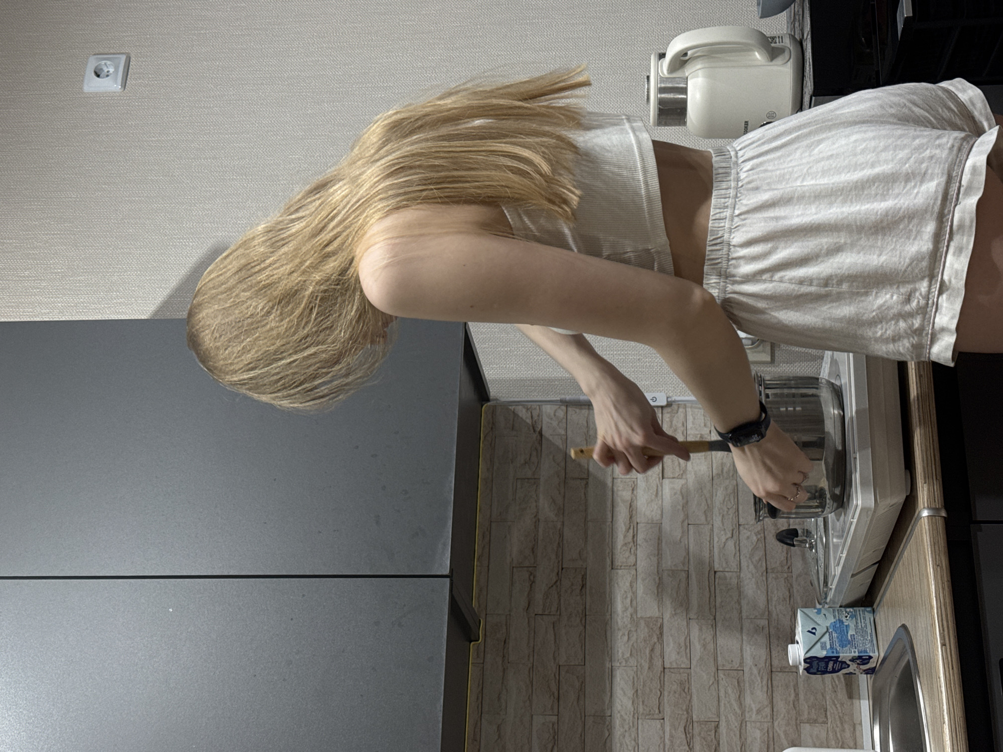 A photograph shows a young woman with long blonde hair wearing a white top and shorts stirring a pot on a white stove in a kitchen. The kitchen has gray cabinets, a beige brick-patterned backsplash, and a white rice cooker is visible. A white electrical outlet is on the wall. A carton of juice sits near the sink. The location is in Perm, Russia.