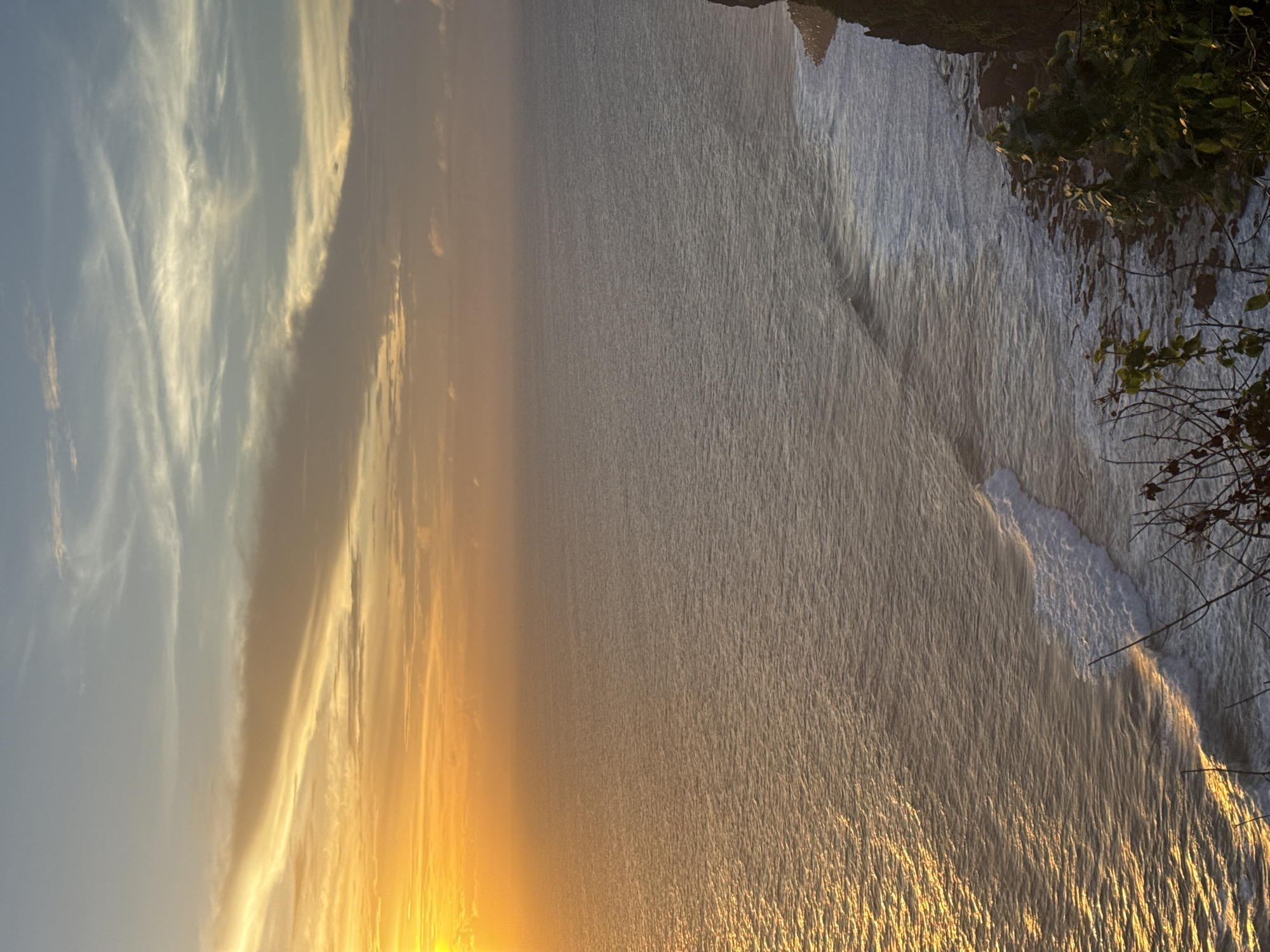A photograph of a sunset over the ocean. The sun is low on the horizon, casting a golden light over the water and the sky. The sky is filled with clouds, some of which are illuminated by the setting sun. The ocean is calm, with small waves lapping at the shore. There is some vegetation visible in the upper right corner of the image. Location: Unknown, Unknown.