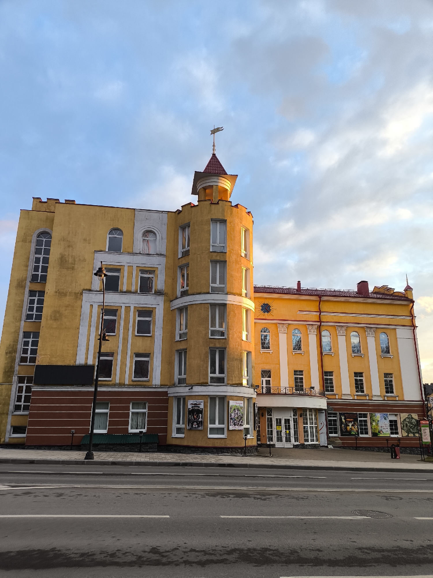 The image shows a yellow building with a prominent tower located in Smolensk, Russia. The building has a mix of architectural styles, with elements resembling a castle tower and more traditional windows and columns. The facade is yellow with white and brown accents. The tower has a red roof and a weather vane. A road runs in front of the building. The sky is cloudy with hints of blue. There are no people visible. Posters are displayed on the building's lower levels. A few streetlights are present.