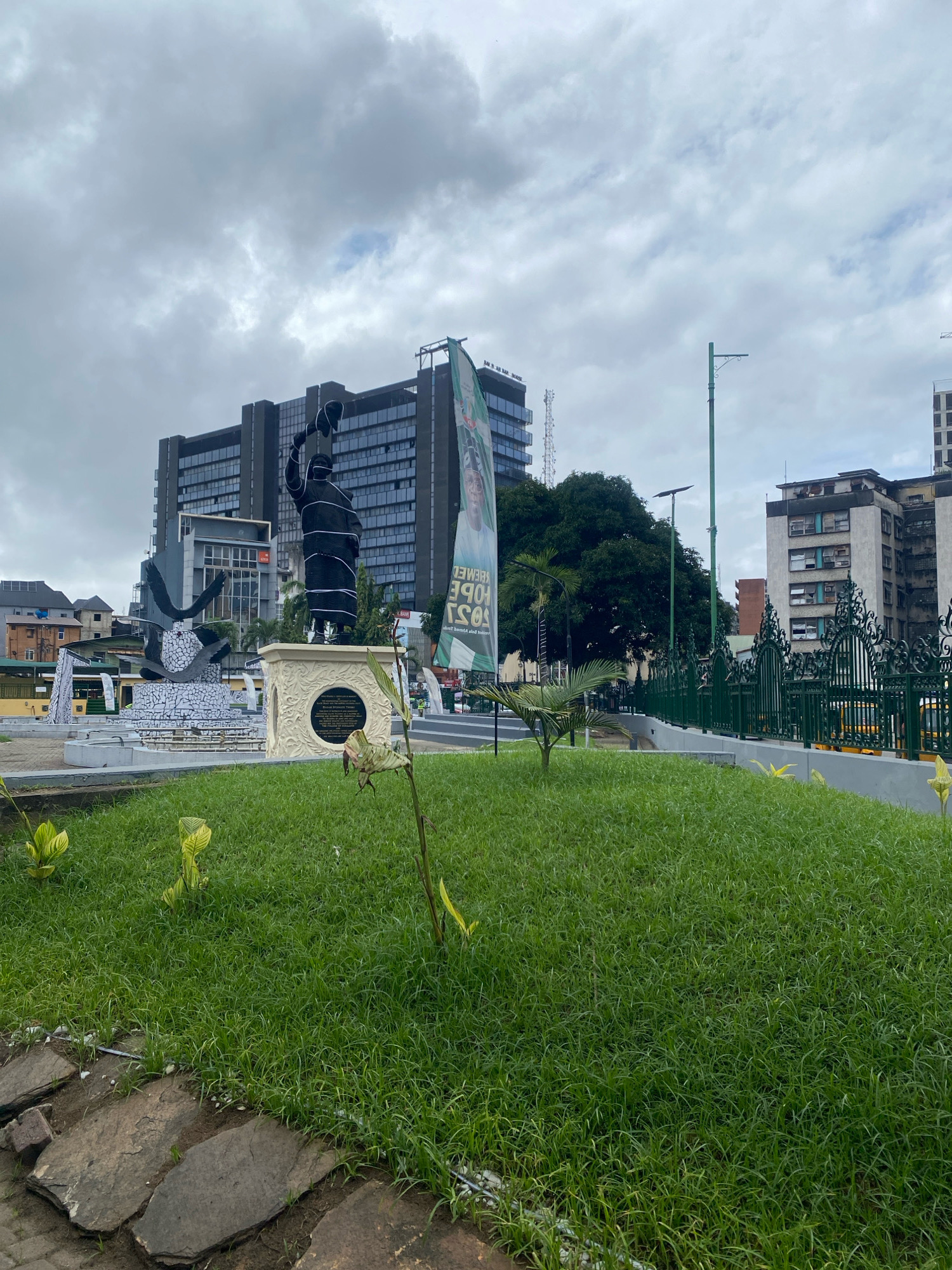 The image captures a public square on Lagos Island, Nigeria, under a cloudy sky. A prominent statue of a standing figure holding a hat is visible, situated on a decorative pedestal. To the left of this statue, another sculptural element, seemingly abstract and made of mosaic tiles, is present. In the background, tall buildings with modern architecture dominate the skyline, characterized by dark glass facades. A large green banner with text and a portrait is unfurled, attached to a tall structure. The foreground features a well-maintained lawn with scattered small plants and a pathway made of stone slabs. A decorative green metal fence runs along the middle ground, separating the lawn from a street with yellow vehicles visible behind it. The environment appears to be a public urban space, possibly a monument or a civic center, with a mix of natural elements like grass and trees alongside constructed architecture.
