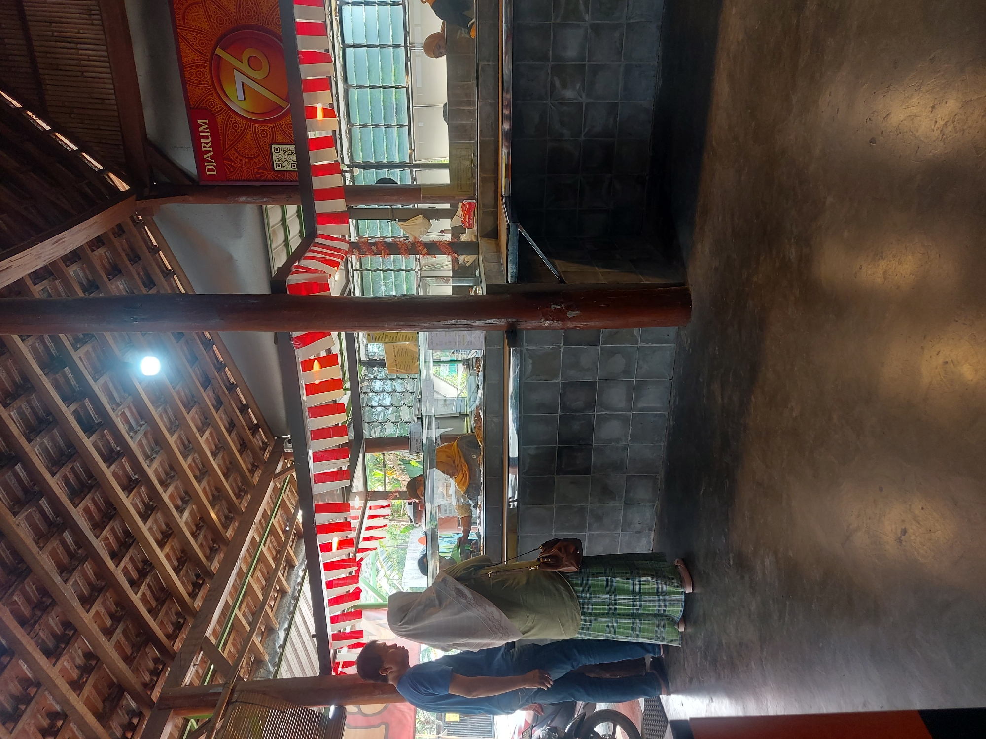 This image depicts an outdoor food stall setting in Kricak, Indonesia. The stall features a wooden-beamed, tiled roof structure with hanging red and white banners. In the foreground, a man in a blue t-shirt and jeans stands next to a woman wearing a green hijab and a checkered green skirt. They are looking towards a food display counter. Behind them, a motorcycle is partially visible. The food counter is made of tiled material and has glass enclosures displaying food items. Several individuals appear to be working behind the counter, including a woman in a yellow hijab. The ground is a polished concrete surface. A large banner with the word "DJARUM" and the number "76" in a circular emblem is visible on the back wall, suggesting a promotional event or anniversary. The environment appears to be a warm, possibly humid, day, with natural light filtering through the open sides of the structure.