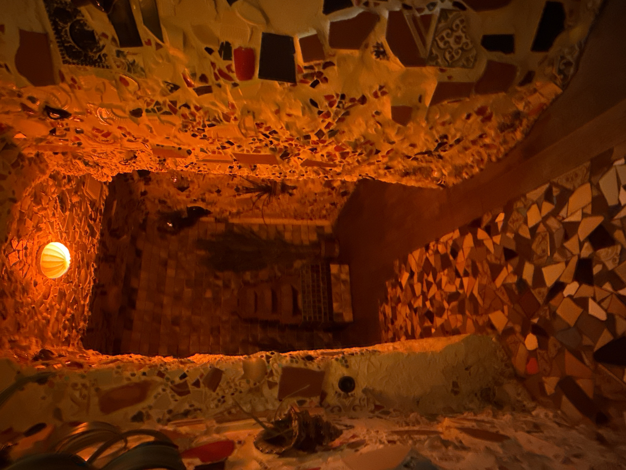 The image depicts an interior space in Moscow, Russia, decorated with intricate mosaic work. The walls and ceiling are covered with a variety of tiles and fragments, creating a textured and colorful surface. A light source illuminates the left wall, casting a warm glow and highlighting the mosaic details. The space appears to be a grotto or a small chamber, possibly part of a larger architectural structure. The scene has an artistic and somewhat surreal quality due to the elaborate mosaic design. There are miscellaneous objects scattered in the foreground, seemingly part of the mosaic construction process or debris. The view is from a low angle, looking upwards into the interior.