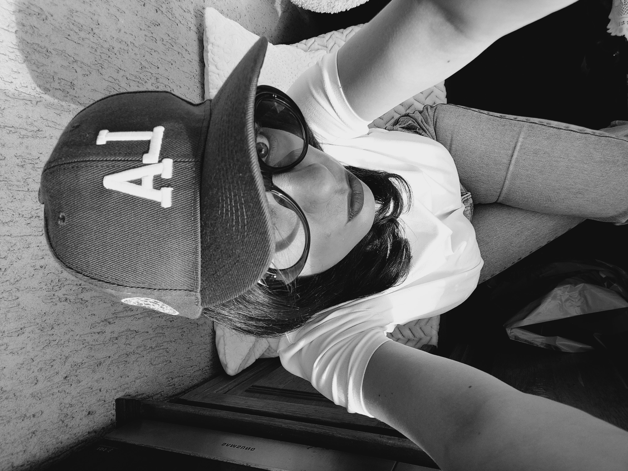 A black and white, high-angle, wide shot shows a person wearing a baseball cap and glasses. The person is facing forward and looking directly at the camera. They are wearing a white t-shirt and jeans. The background consists of a wall with a textured pattern and some bedding. The person's arms are extended towards the camera, as if taking a selfie. The scene is indoors. The location in the city of Sofia, Bulgaria is not discernable from the image.