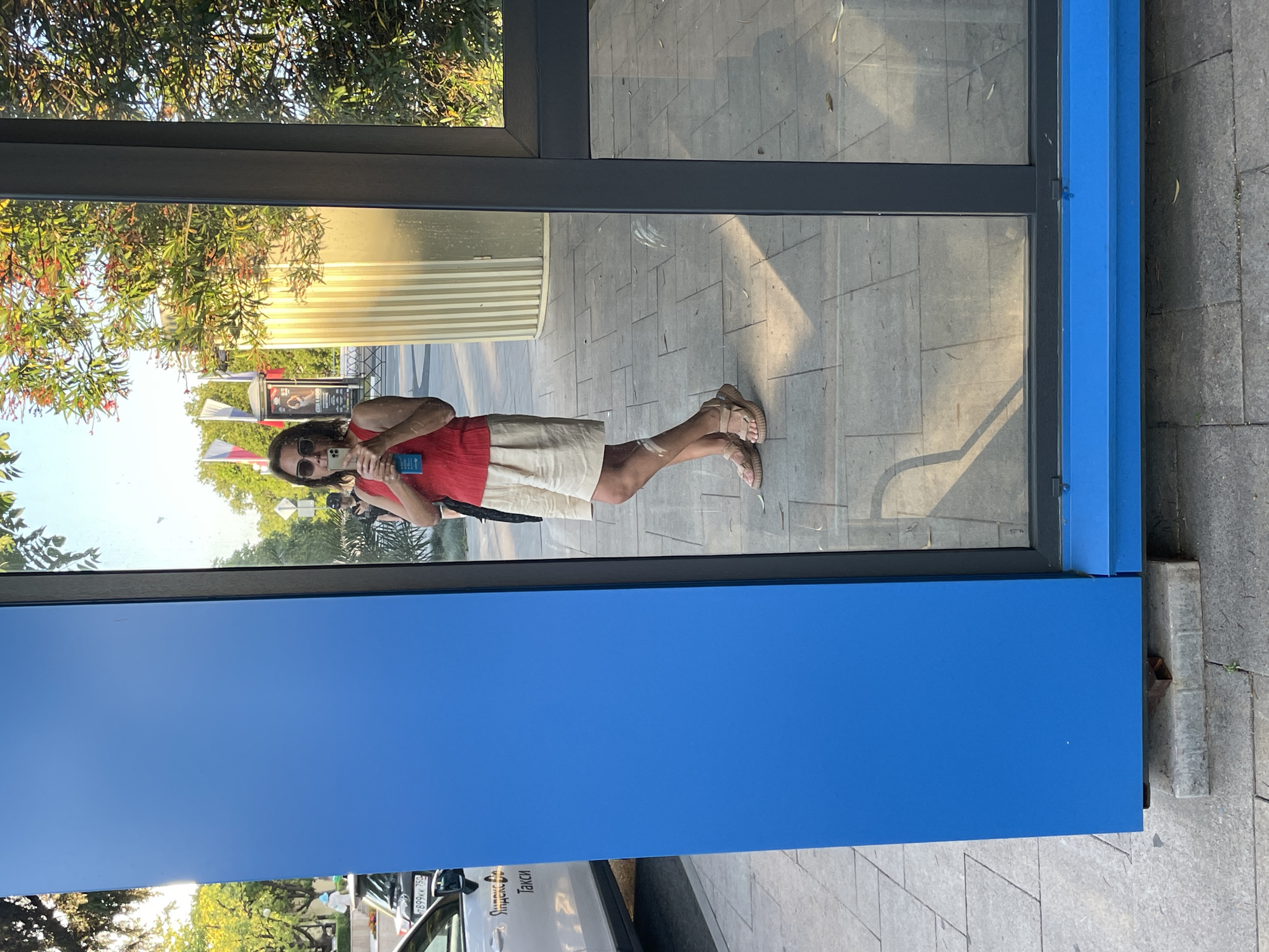 A person is taking a selfie in a reflection of a glass structure. The person is wearing sunglasses, a red top, and beige shorts. They are holding a blue object, possibly a phone. The reflection shows a paved area with rectangular stones, some greenery, and buildings in the background. The time of day appears to be daytime, with sunlight casting shadows. The overall mood seems casual and relaxed. There is visible text on a white car in the reflection, which says "Яндекс Такси" (Yandex Taxi).