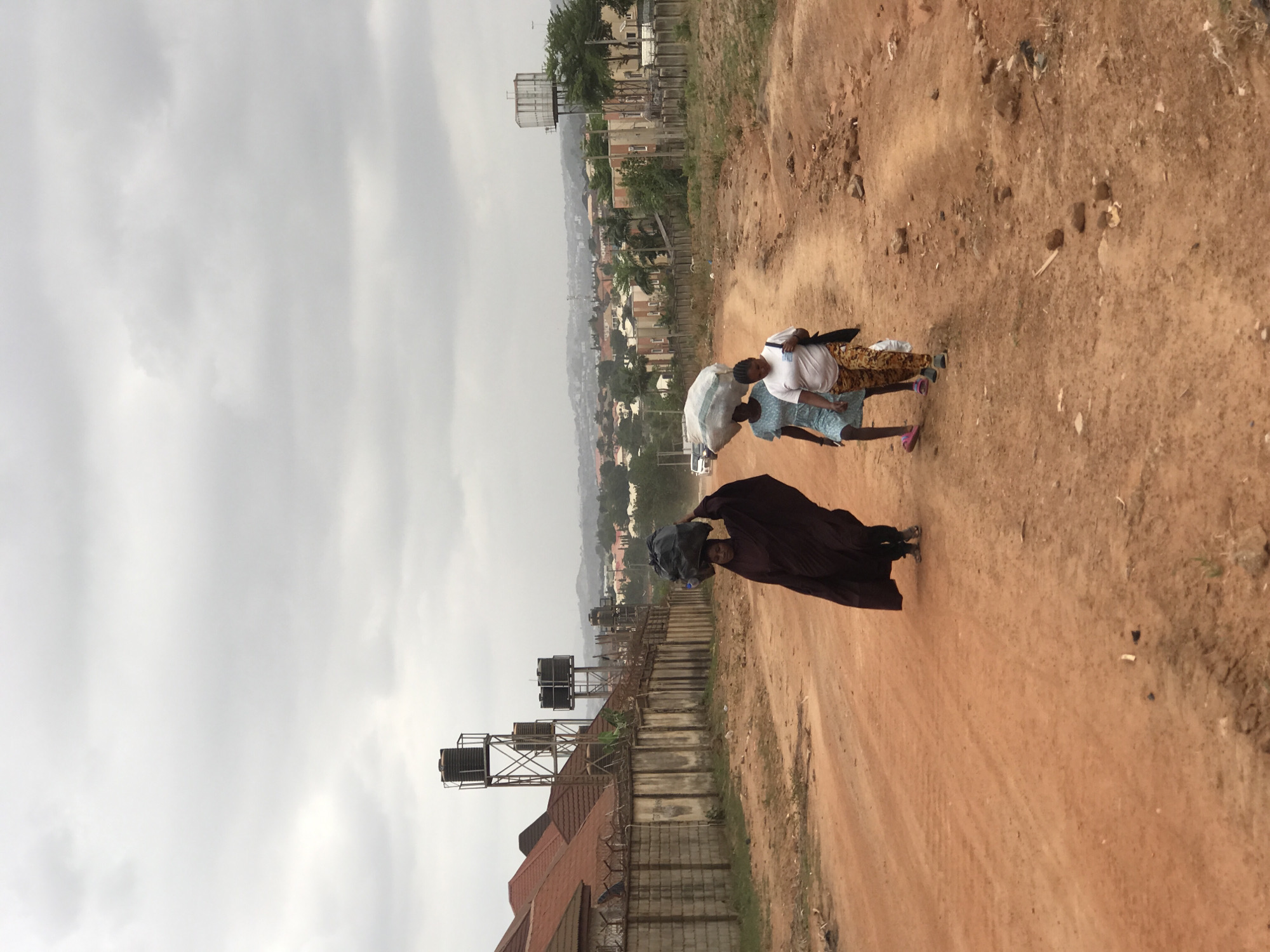 The photo depicts an unpaved, red dirt road in what appears to be a residential area of Abuja, Nigeria, evidenced by the presence of buildings and water towers in the background. Four individuals are visible on the road. The foremost figure is wearing a dark, draped garment and carrying a dark item on their head. Behind them, three individuals are walking together, one carrying a light-colored sack or bag. The sky is overcast, and the general environment suggests a developing urban setting. A structure with a red roof and multiple water tanks is visible on the left side of the road.