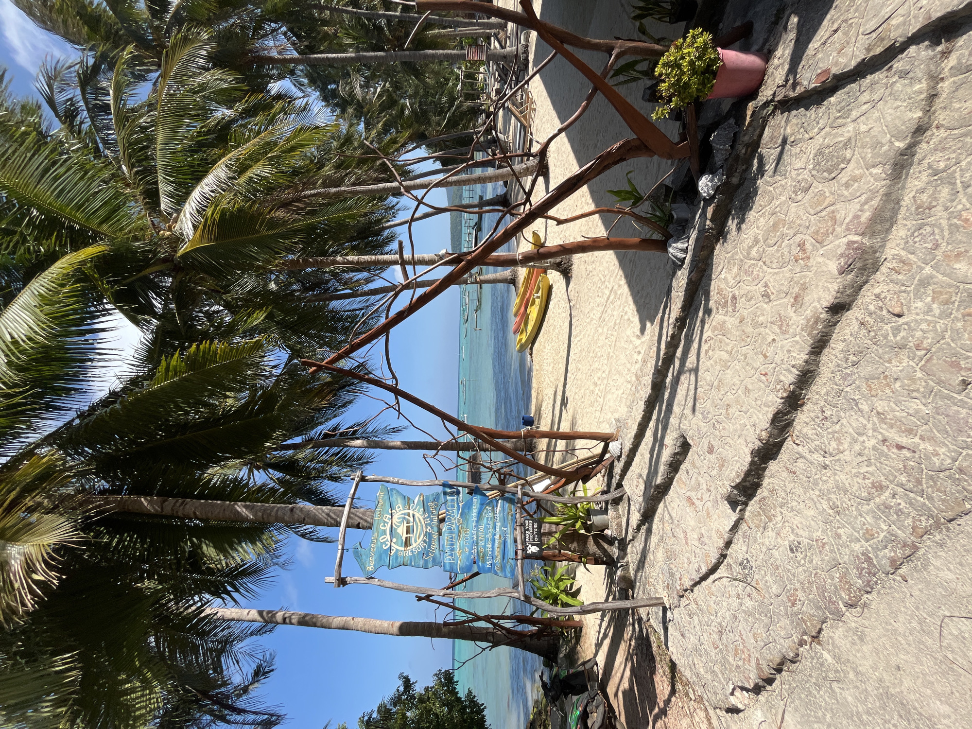 In Legonipah, Indonesia, a beachside resort named Go Casa Resort is depicted. The scene is set under a bright, clear sky with lush green palm trees lining the sandy shore. In the foreground, a paved walkway leads to the beach, with a signpost in the center providing information about the resort's amenities, including food, rentals for motorbikes, bicycles, and boats, as well as swimming and snorkeling spots. Two yellow kayaks are visible on the sand, hinting at water activities. In the background, the turquoise ocean stretches towards the horizon, dotted with small boats. The beach is furnished with lounge chairs, suggesting a relaxing atmosphere. A motorcycle is parked to the left of the signpost.