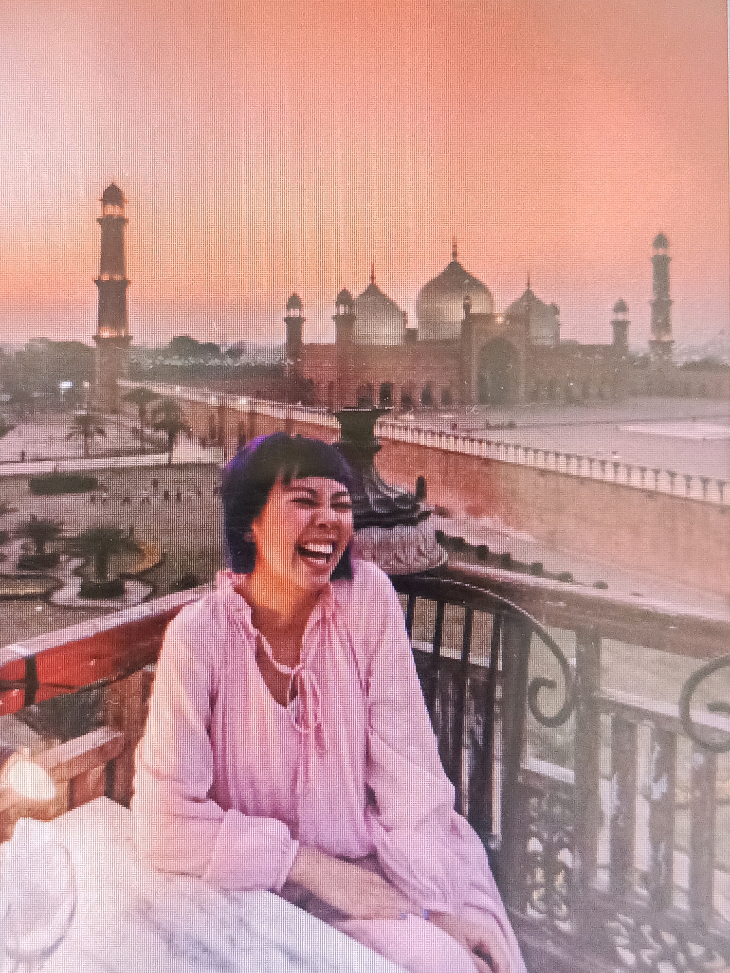 The image shows a woman with short, dark hair and a purple hue, laughing with her mouth wide open and eyes closed. She is wearing a pink, long-sleeved shirt with a ruffled neckline and tie. She is seated at a table with a white tablecloth, with a dark railing in the foreground. In the background, a large, ornate building with domes and minarets is visible against a soft orange sky, suggesting sunset or sunrise. The building appears to be a mosque or palace, and the overall scene depicts an outdoor setting with a garden and walkway. The location is identified as Rasoolpur, Pakistan, and the building resembles the Badshahi Mosque in Lahore.