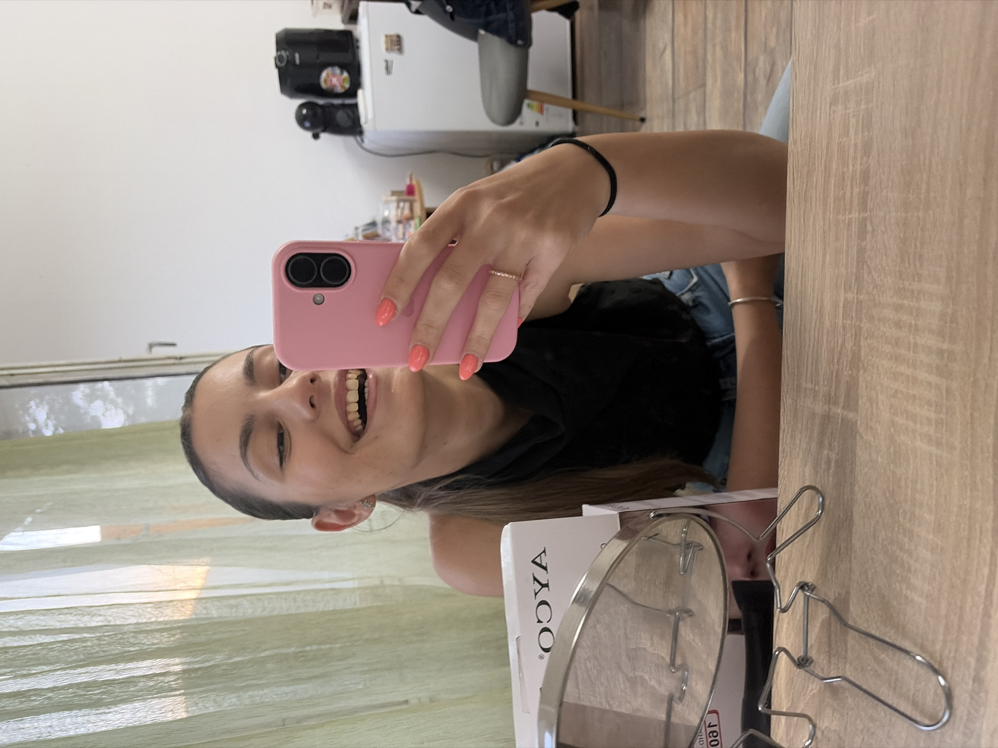 A woman is taking a selfie with a pink smartphone. She is smiling and facing the mirror with green curtains. There is a wooden table to the right with a kitchen lid holder on top. In the background, there is a refrigerator, a stool, and some kitchen appliances. The location may be in Sofia, Bulgaria, given the available information.