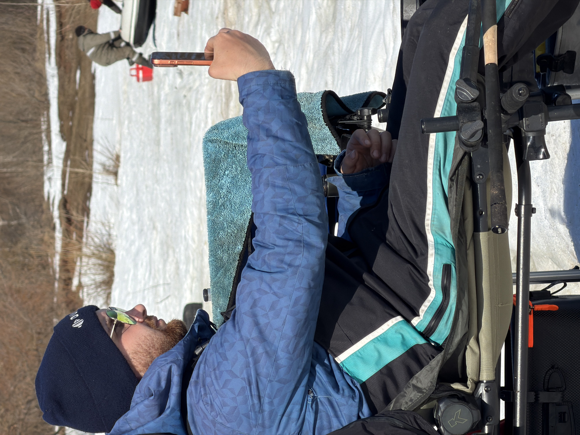 A person wearing a blue and black jacket, dark beanie, and sunglasses is sitting outdoors, holding a smartphone, with what appear to be fishing rods and tackle next to them. The ground is covered in snow, and in the background, another person is on a sled. The environment suggests a cold, possibly rural or semi-rural location. There are trees without leaves in the distance. The location is Noviy, Russia.