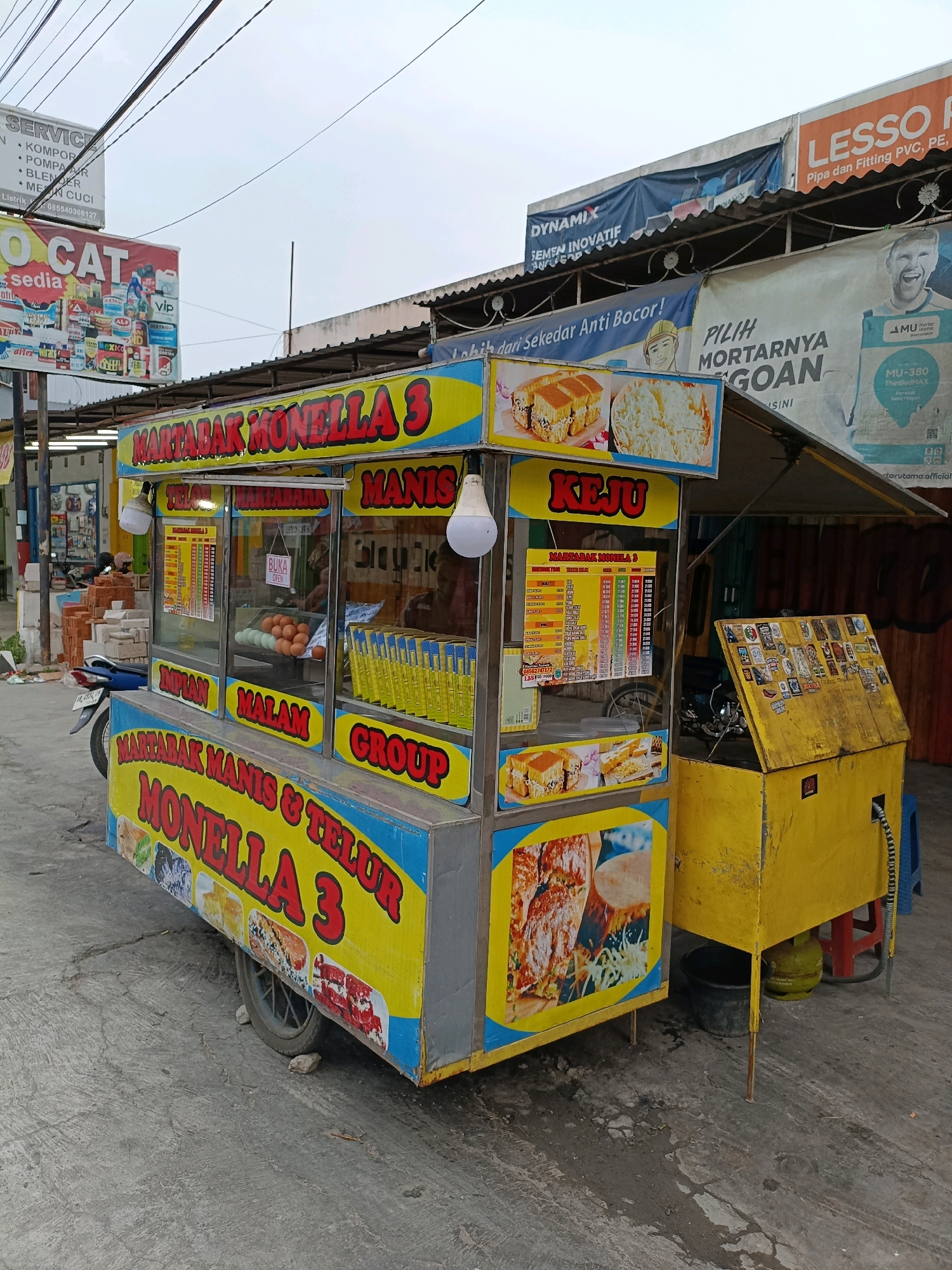 An outdoor food cart for "MARTABAK MANIS & TELUR MONELLA 3" is parked on a paved surface, likely a street or sidewalk, in Maguwoharjo, Indonesia. The cart is predominantly yellow and blue, with red lettering. Various advertisements are visible in the background, including "LESSO Pipa dan Fitting PVC, PE, PP-R" and "DYNAMIX SEMEN INOVATIF". The cart itself displays images of martabak and a menu board. Inside the cart's display window, eggs are visible alongside bottles of yellow liquid. A blue motorcycle is parked to the left of the cart. To the right, a yellow chest with a propane tank underneath sits beside the cart. The sky is overcast.
