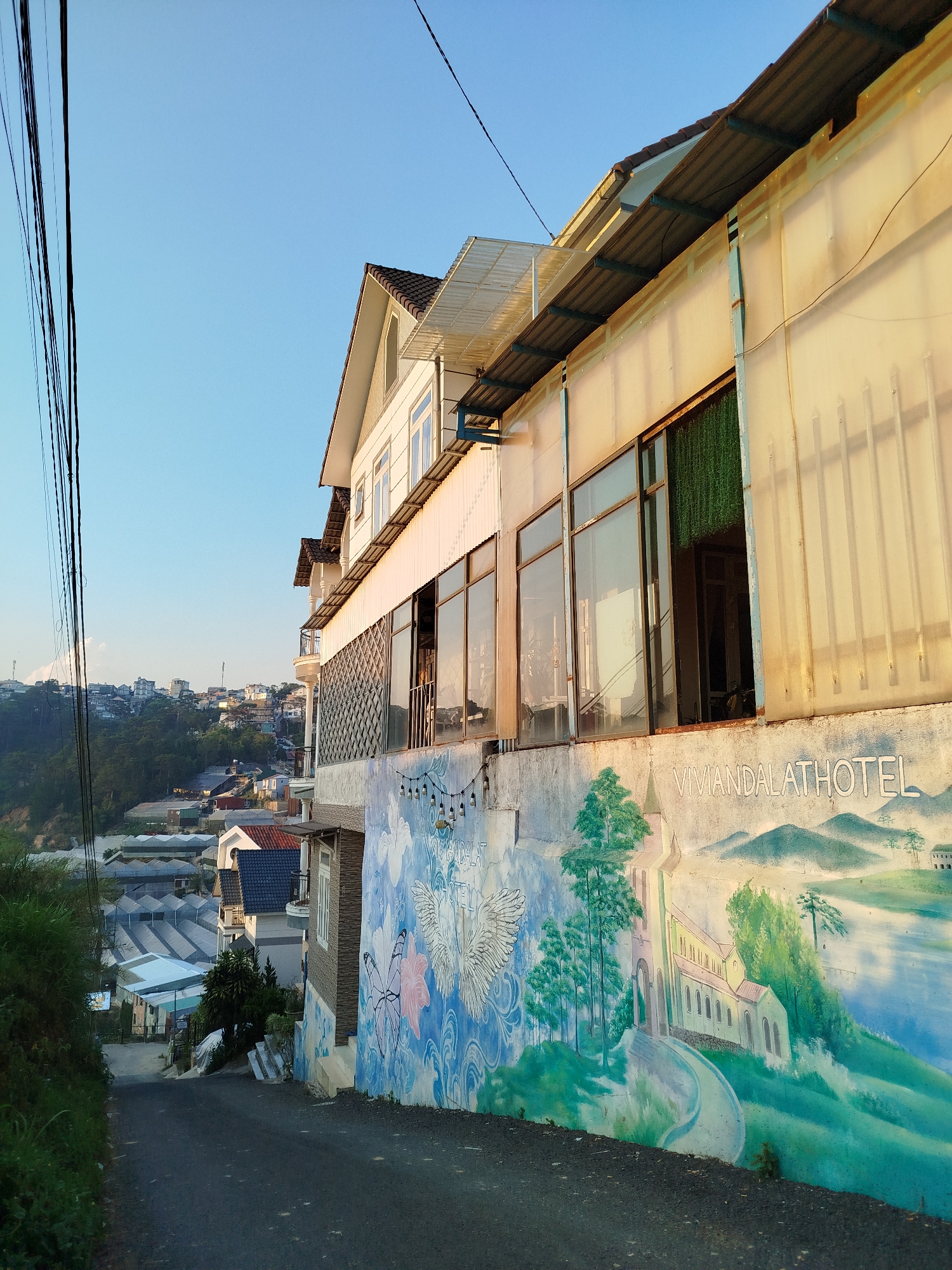 Captured from a low angle on a paved road, the image features a building with an aged, light-colored exterior. A mural covers the building's lower section, depicting a landscape with trees, a church, and distant mountains, with the words "VIVIANDALATHOTEL" painted above. The building features large windows, some with visible reflections, and awnings overhead. A row of string lights hangs along the top of the mural.
The road leads downhill, revealing a view of Da Lat, Vietnam, characterized by a mix of residential buildings and greenhouses. In the distance, a hazy skyline is visible under a light blue sky. Utility lines stretch across the upper portion of the frame. The perspective suggests the photographer is positioned on a road running alongside the building.