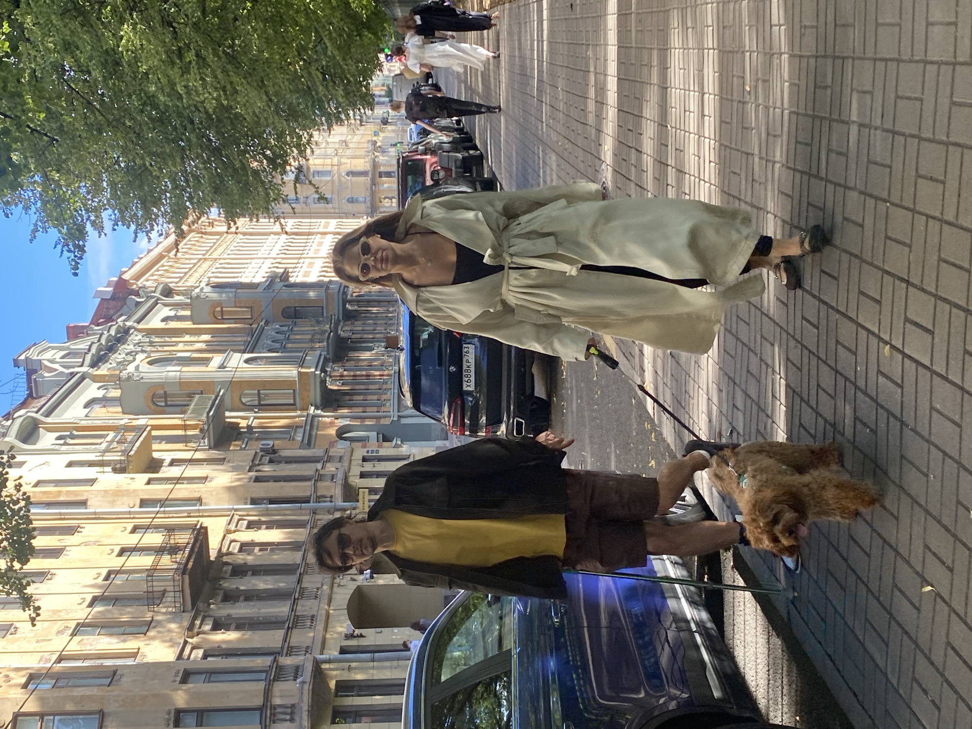A man and a woman are walking on a sidewalk in Saint Petersburg, Russia, with a small, fluffy brown dog on a leash. The man, wearing sunglasses, a yellow t-shirt, and brown shorts, walks ahead of the dog, holding a walking stick in his left hand. The woman, also wearing sunglasses, is in a light-colored trench coat and dark pants, walking beside and slightly behind the dog. Behind them, a group of people, including a woman in a white dress, are walking further down the sidewalk. The buildings lining the street are ornate and appear to be historic architecture, typical of Saint Petersburg. Trees with green foliage provide shade on the right side of the sidewalk. Cars are parked along the street. The sun is shining, casting shadows on the paved sidewalk.