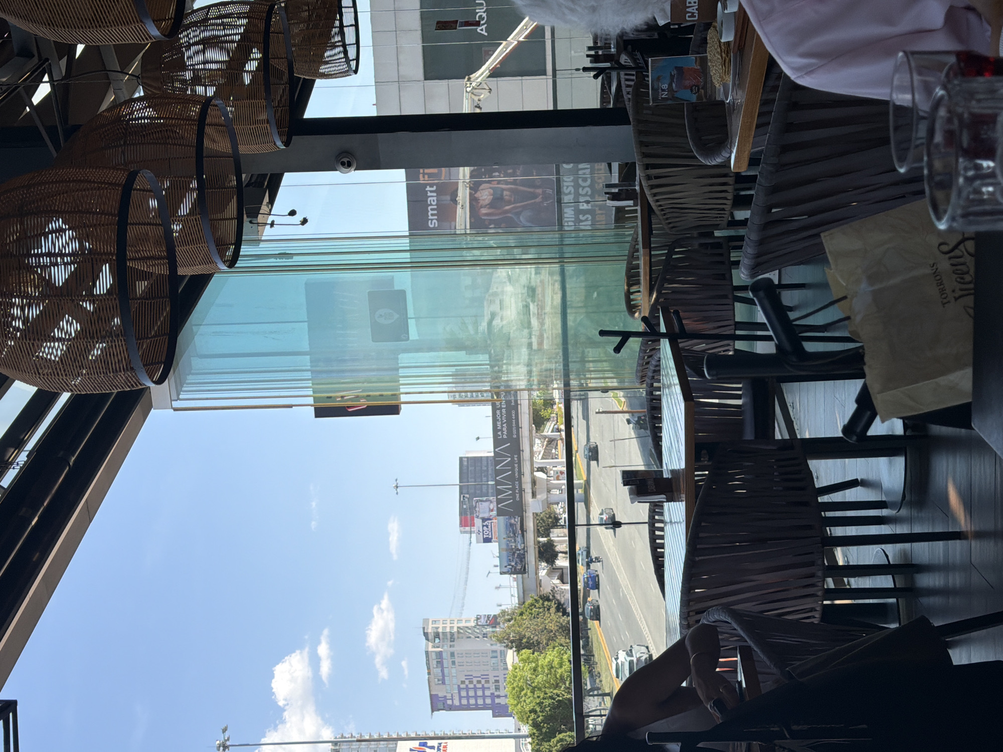 This image offers a vibrant view from inside a modern restaurant or cafe, looking out through large glass windows onto a sunny cityscape. The interior features contemporary design elements, including several light brown wicker chairs with dark metal frames around tables. Decorative, elongated wicker-like light fixtures hang from the ceiling, creating an inviting ambiance. On one of the tables, clear drinking glasses, a small dish, and a book or magazine labeled "N.8" are visible. A paper bag displaying "TORRONS Vicens" suggests the presence of snacks or desserts. Parts of patrons, including an arm with a bracelet and another arm with a pink shirt, indicate people are present, likely enjoying a meal or conversation.
Outside, the scene is bathed in bright daylight under a clear blue sky with scattered white clouds. A multi-lane road parallels the building, busy with cars and trucks. The urban landscape includes numerous multi-story buildings, some featuring unique architectural designs like purple-squared facades, and interspersed green trees. Prominently displayed are several large billboards. One advertises "smart fi" alongside a person exercising, with text suggesting a "GIMNASIO CON PISCINA" (Gym with Pool). Another billboard for "AMANA ONE PLACE - UNIQUE LIFE" explicitly states "LA MEJOR UBICACIÓN PARA VIVIR EN PUEBLA" (The Best Location to Live in Puebla) and provides a phone number: 221 644 4400. This confirms the location as Puebla, Mexico. The overall impression is a comfortable, well-lit establishment offering panoramic views of a lively urban environment.