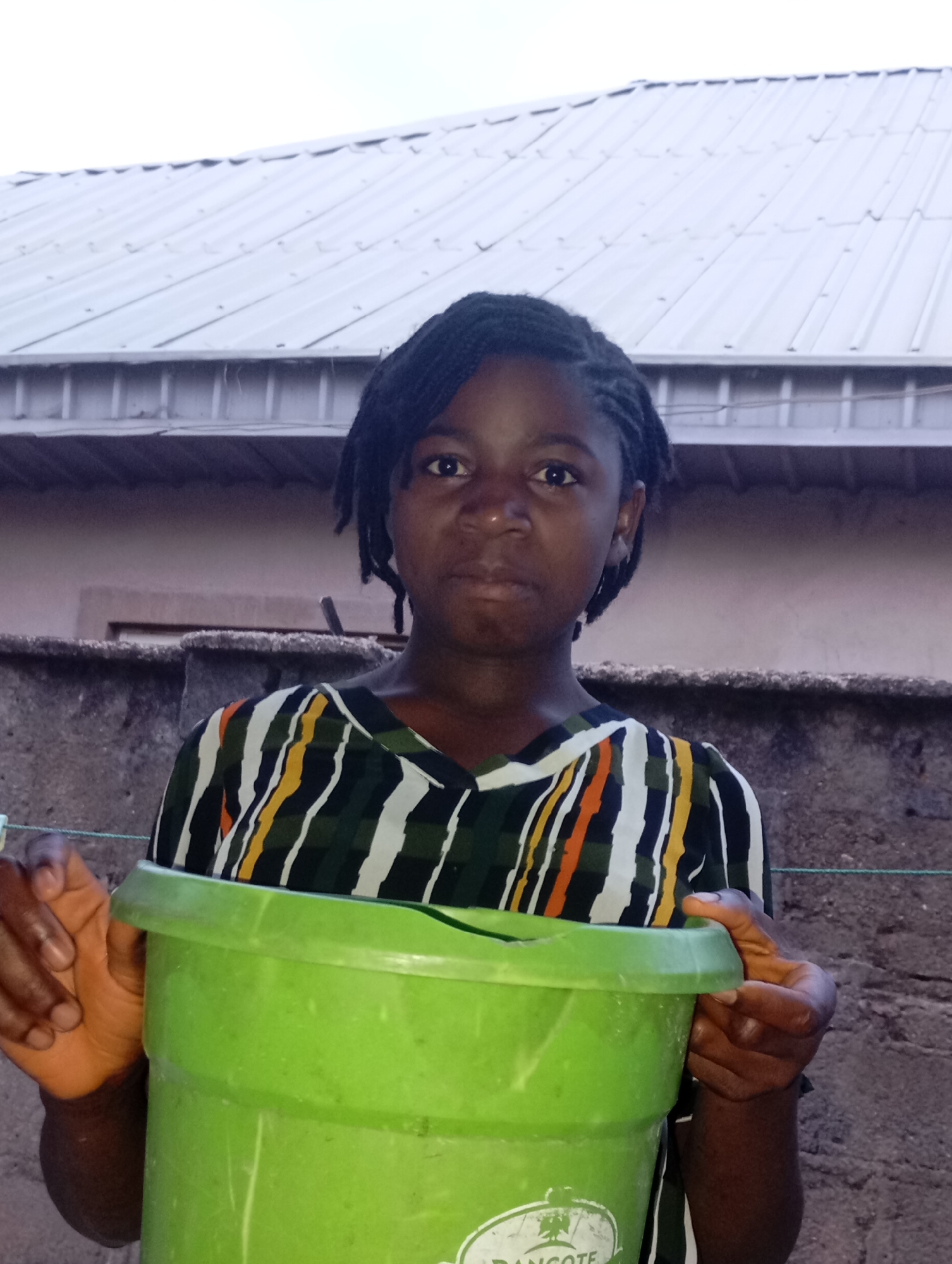 A young girl with dark skin and braided hair stands outdoors in Jos, Nigeria, holding a large, light green plastic bucket. She wears a v-neck shirt with a pattern of vertical and diagonal stripes in green, white, orange, and black. Her expression is neutral as she looks directly at the camera.
She holds the rim of the bucket with both hands, positioned in front of her. In the background, there is a low, rough-textured concrete or brick wall. Behind the wall, the roof of a building with light-colored corrugated iron sheets is visible against an overcast sky, suggesting daytime. A thin green line, possibly a clothesline, stretches horizontally behind the girl's right arm.
The brand name "DANGOTE" with a logo is visible in white text on the lower front of the green bucket. The overall setting appears to be a residential or domestic outdoor area.