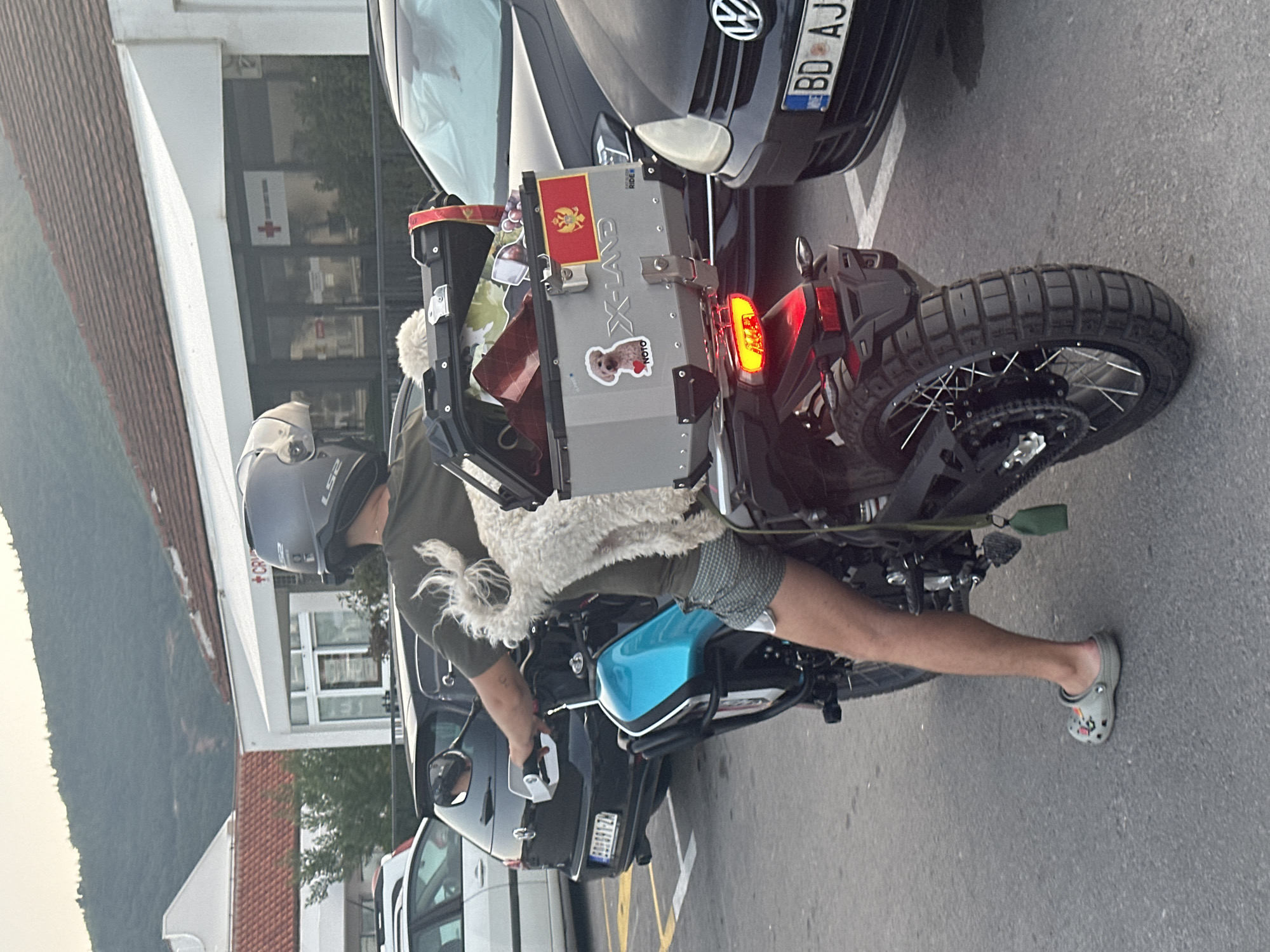 A person is riding a motorcycle in Budva, Montenegro, with a small white dog perched on the back of the motorcycle. The person is wearing a dark green t-shirt, checkered shorts, and grey Crocs. They have a dark grey helmet on their head. The motorcycle has a large silver storage box on the back, adorned with a Montenegrin flag and a sticker of a dog. The dog is white and fluffy, with its head facing forward. The scene takes place in a parking lot, with several cars parked nearby. In the background, there is a building with a red cross sign, suggesting it might be a medical facility, and a wooded hill. The overall lighting indicates it is either early morning or late afternoon.