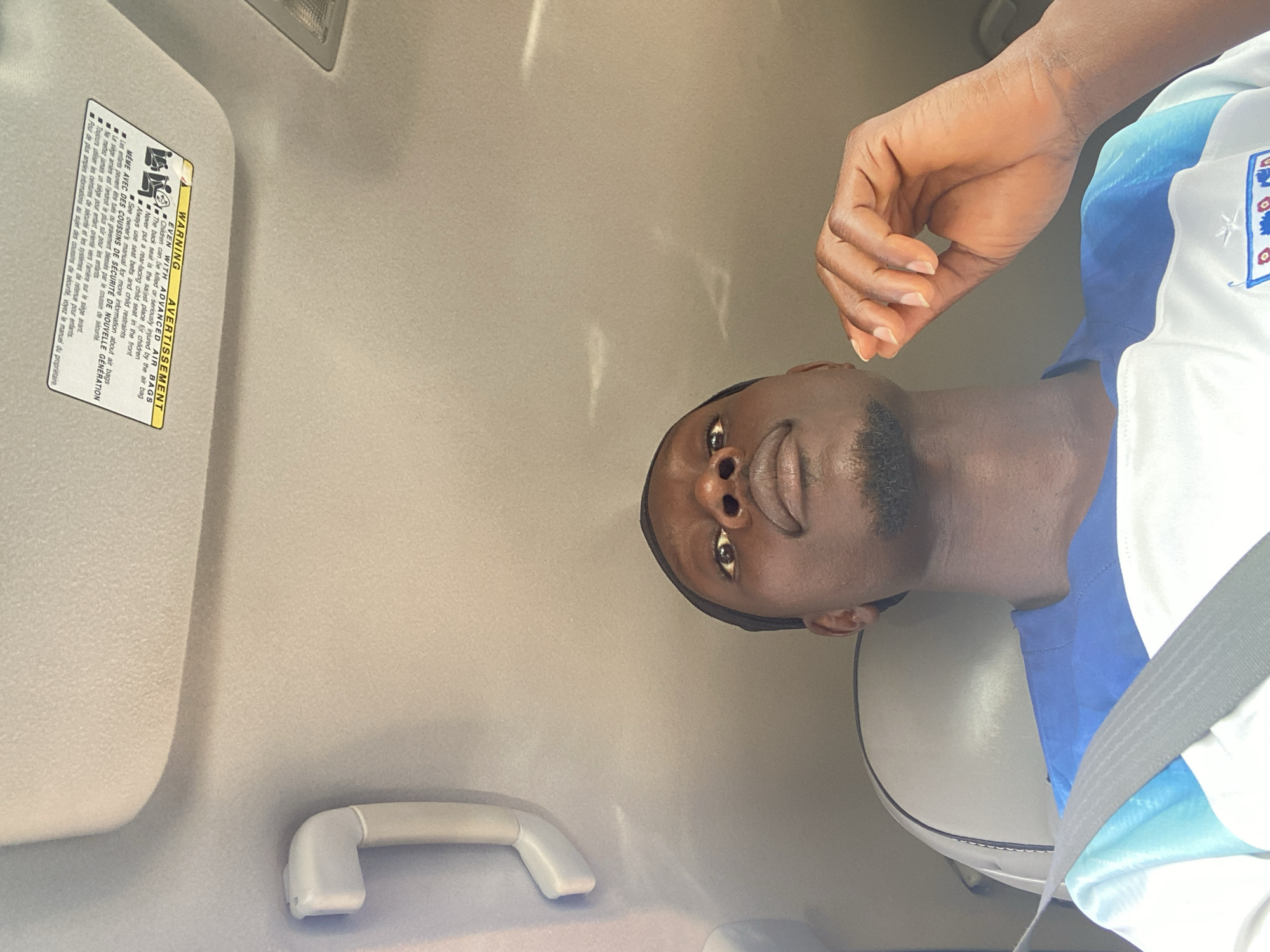 The image shows a close-up, low-angle shot of a Black man wearing a blue and white jersey, seated inside a vehicle. He is looking upwards, with his right hand resting on his chin and his fingers extended. A seatbelt is visible across his chest. The interior of the car is dimly lit, with a visible overhead handle and a warning label for airbags attached to the car's ceiling. The background consists of the car's headliner and a sliver of a light fixture. The man has a short beard and is wearing a dark skullcap.