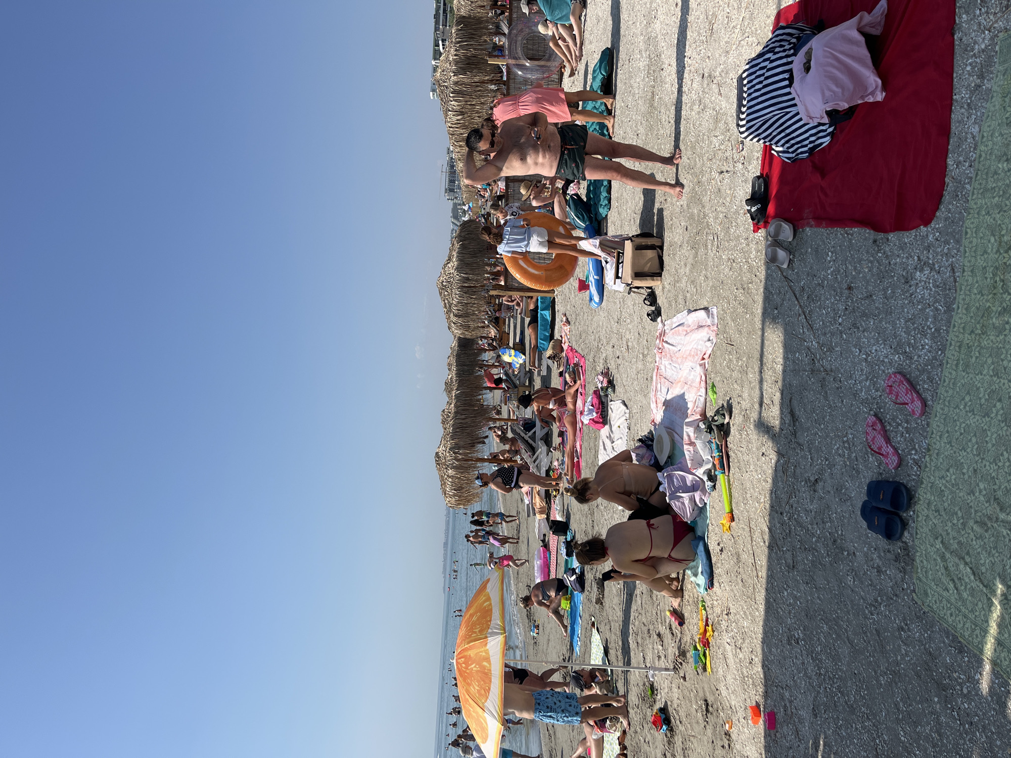 The image depicts a crowded beach scene in Năvodari, Romania, on a clear, sunny day. Numerous people are sunbathing, relaxing on towels and lounge chairs, and swimming in the calm sea. Several umbrellas, including a large orange one in the foreground, provide shade. Beach toys, inflatable rings, and bags are scattered on the sand. In the background, there are rows of thatched umbrellas, beach structures, and buildings along the coastline. The overall atmosphere is one of leisure and recreation.