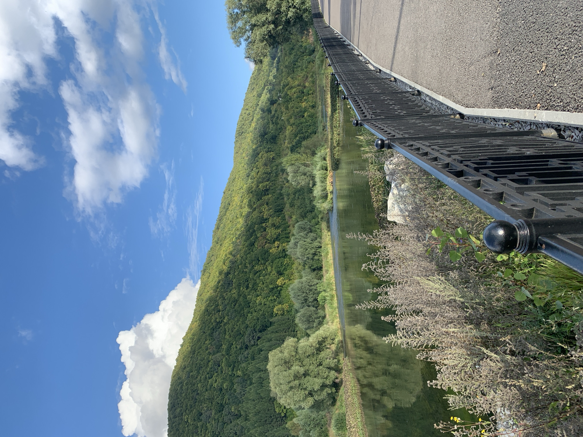A paved walkway with a black metal railing runs along the edge of a wide, calm river. The river is bordered by lush green trees and a steep, tree-covered hillside. The sky is blue with scattered white clouds. The scene is peaceful and scenic, suggesting a park or recreational area. This location is the embankment of the Ufa River in the city of Ufa, Bashkortostan, Russia.