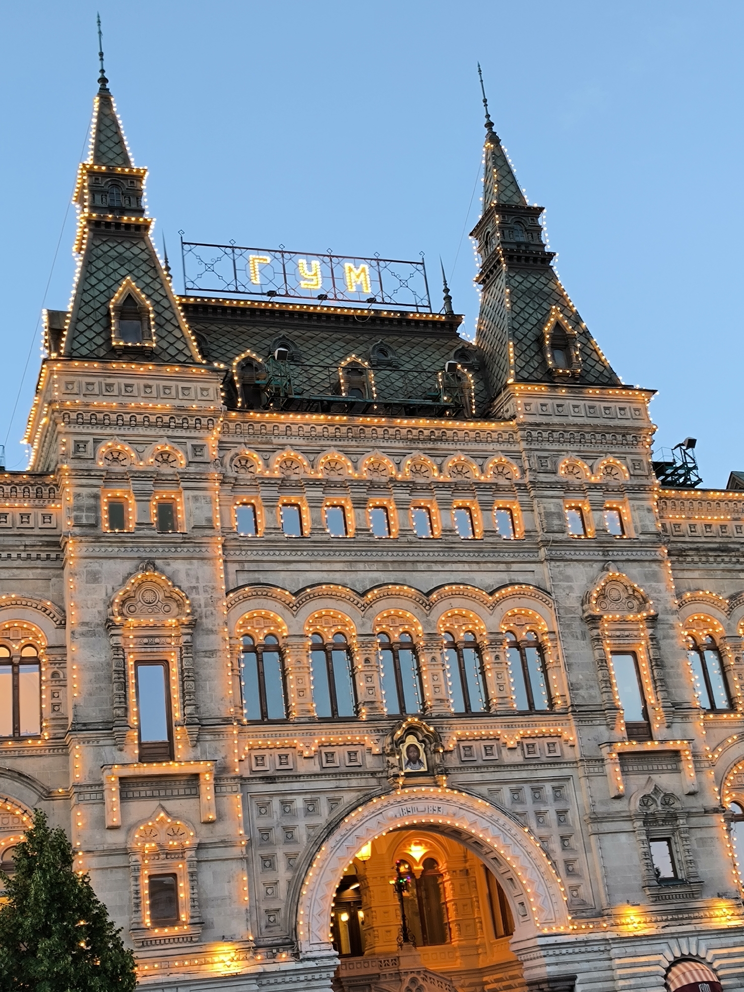 The image shows a close-up view of the GUM department store in Moscow, Russia, decorated with festive lights. The architectural style is ornate, with detailed stonework and arched windows. The lights are strung along the building's facade, outlining the rooflines, windows, and decorative elements, creating a warm, inviting glow against the twilight sky. The iconic "ГУМ" sign is visible at the top of the building. There are no people or specific actions depicted in this scene, just the illuminated facade of the historic building. The overall impression is one of grandeur and celebration.