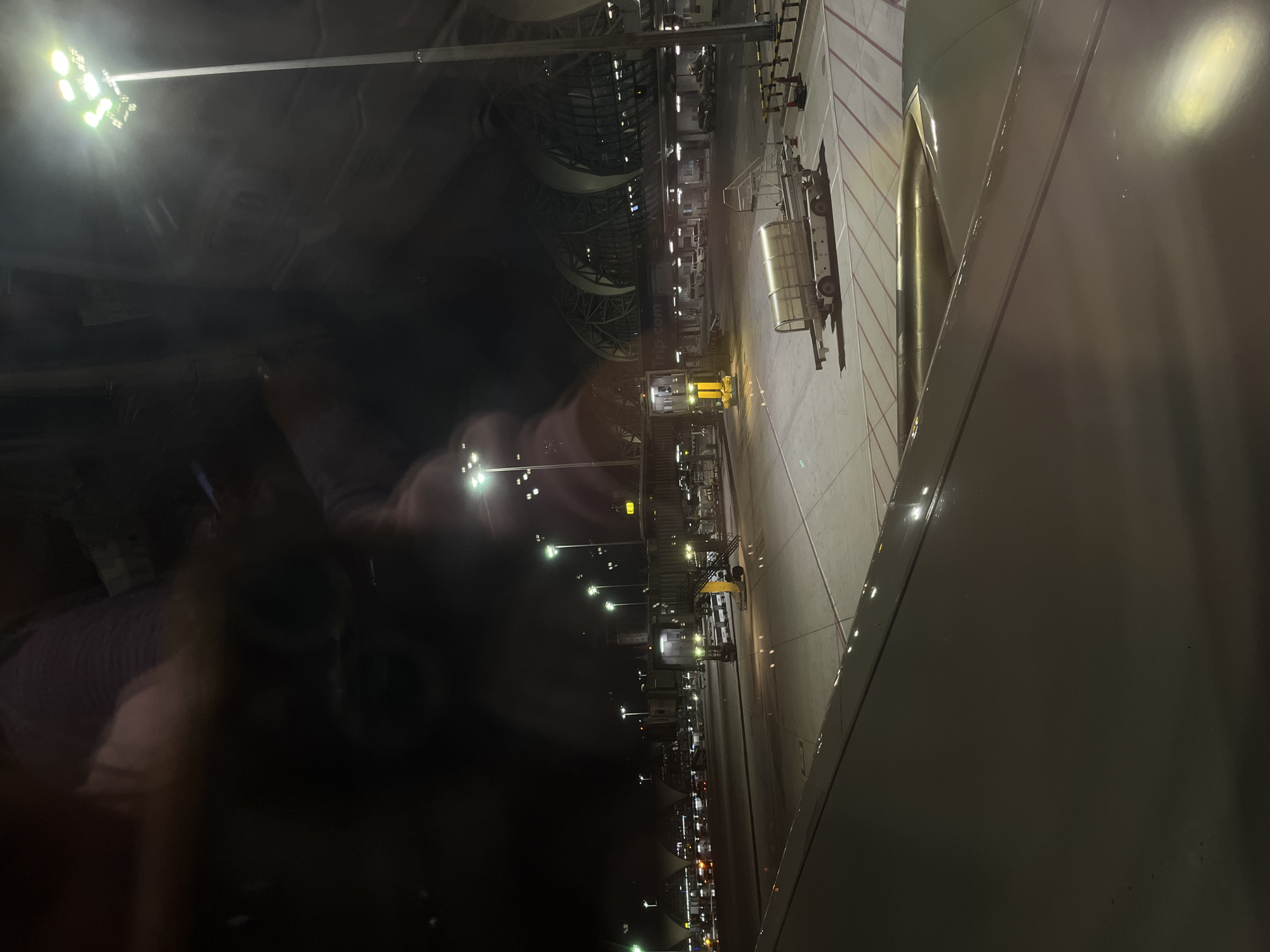 An aerial view at night shows an airport tarmac. A plane's wing is in the foreground, reflecting the tarmac lights. In the mid-ground, a baggage tug is parked near a jet bridge. The background features a large, modern airport terminal building with a distinctive arched roof structure. The scene is illuminated by numerous tall airport floodlights. The location is Suvarnabhumi Airport, located in Nong Prue Subdistrict, Bang Phli District, Samut Prakan Province, Thailand, near Bangkok.