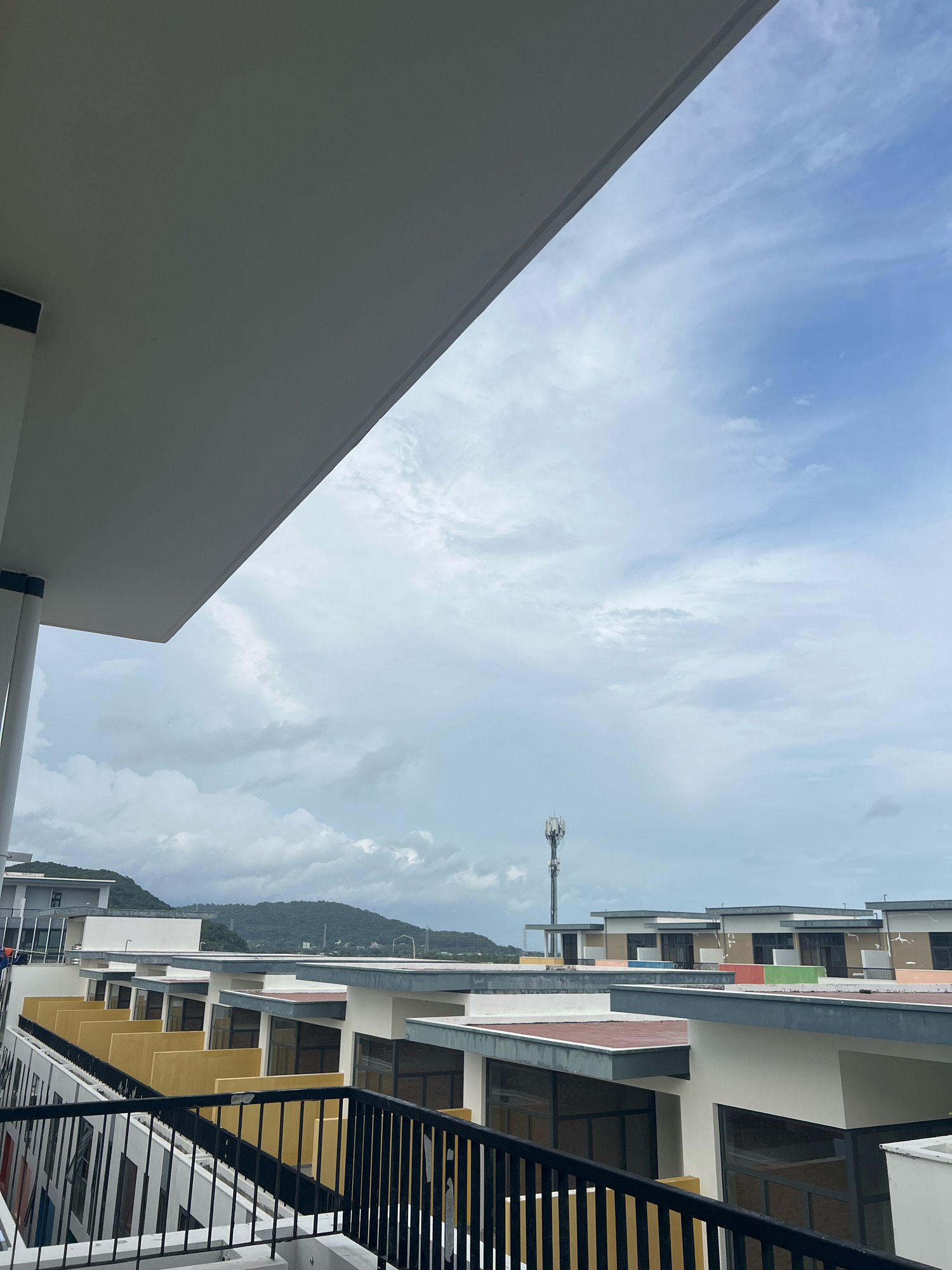 An overcast sky with scattered clouds dominates the upper portion of the image. Below the sky are the rooftops of a modern housing development. The buildings feature white walls, flat roofs with varying colored edges (red, green, blue), and large dark-paned windows. Yellow accent walls are visible on some of the lower sections of the buildings. A black metal railing runs along the foreground, suggesting a balcony or walkway. In the distance, rolling green hills are visible under the cloudy sky, and a tall cell tower stands prominently against the horizon. The scene appears to be in the city of Phú Quốc, Vietnam.