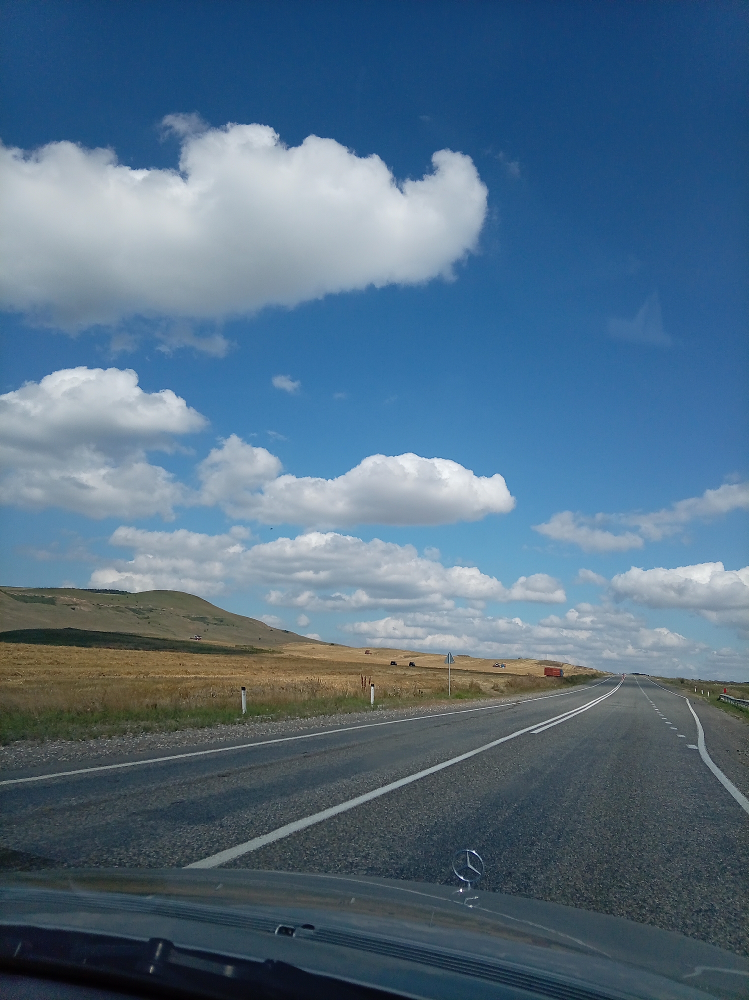 The image captures a view from a car driving on a highway in a rural landscape. The road, paved with asphalt, stretches into the distance, marked by white lines. To the left of the road, a dry, grassy field extends to the base of rolling hills. The hills are covered in sparse vegetation and appear to be a mix of dry grass and some green patches. A few white-topped posts line the side of the road. In the far distance, on the side of the road, a red cargo container is visible. The sky is a vibrant blue, dotted with fluffy white clouds. The foreground shows the hood of a vehicle, with a Mercedes-Benz emblem visible. The scene is devoid of people and animals. The overall impression is one of travel through a vast, open, and somewhat arid countryside.