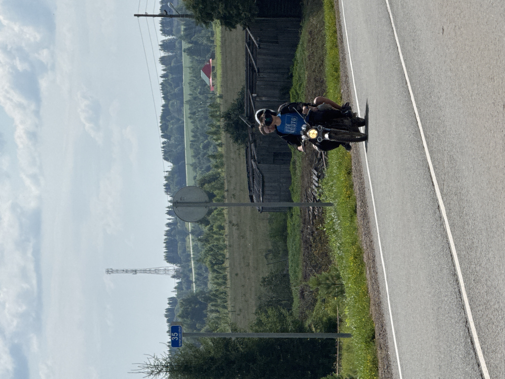 A rural roadside scene features a motorcyclist and a passenger riding a motorcycle on a paved road with white lane markers. The motorcyclist is wearing a black helmet and a blue t-shirt with text that appears to say "VSE KRUTO". The passenger is wearing a white helmet. In the background, there are several dilapidated wooden structures, a green field, a line of trees, and a red and yellow building. A cell tower is visible in the distance. A blue street sign with white text reading "35" and "ПЕРМСКИИ КРАЙ" is posted on a pole to the left of the road. The sky is partly cloudy.