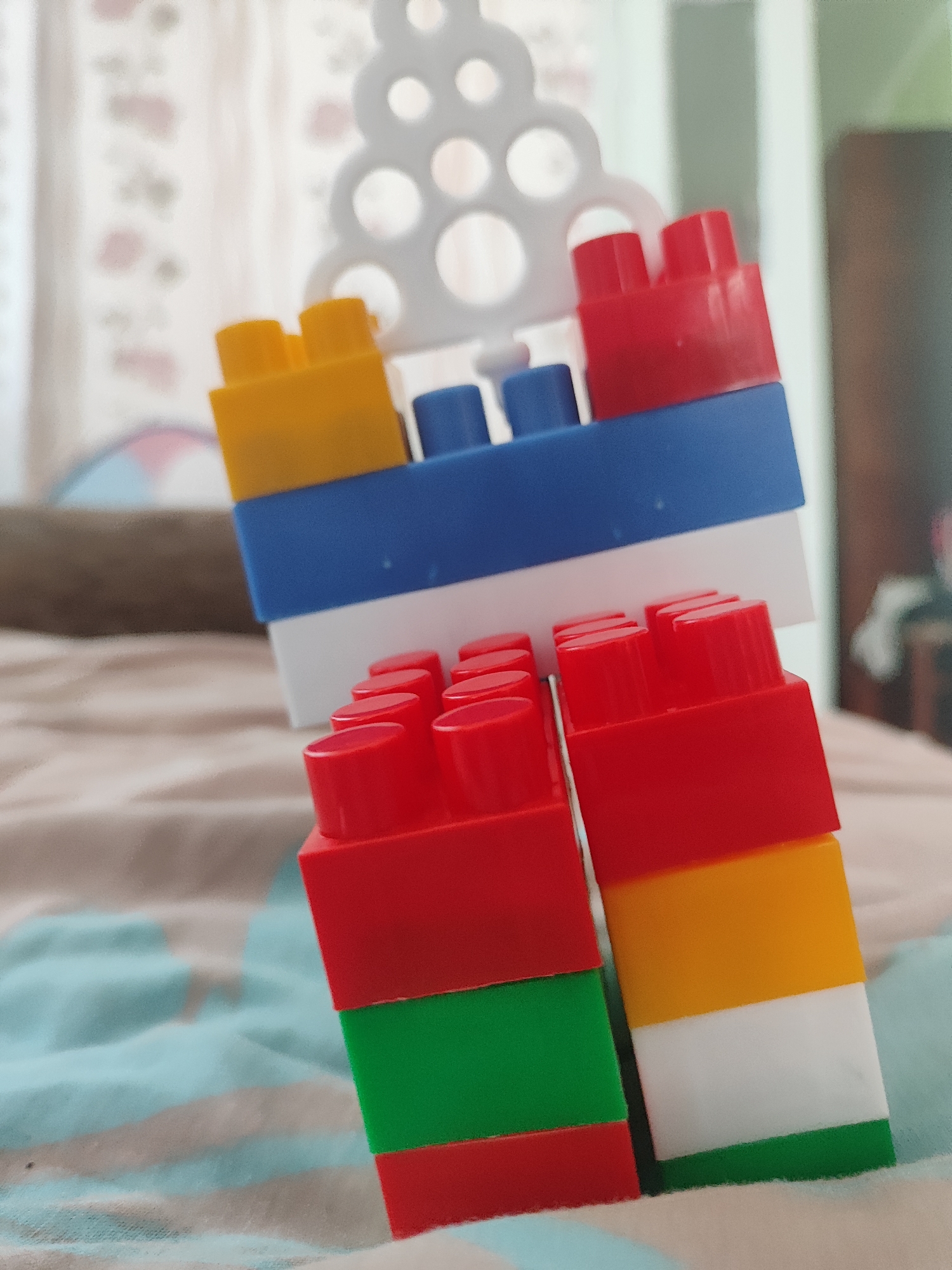 This media file is a close-up, indoor photograph of several colorful building blocks. The subjects are the blocks themselves, arranged in two distinct stacks. One stack, in the background and slightly out of focus, is composed of yellow, blue, and red blocks, with a white, decorative, perforated piece of plastic on top, resembling a large, stylized Christmas tree shape. The second stack, in the foreground and in focus, is made of red, green, red, and white blocks on the left, and red, yellow, white, and green blocks on the right.
The blocks are resting on a soft surface, likely a bed or a blanket, which has a textured pattern of light blue and beige stripes. The lighting suggests it is daytime, with soft, diffused light coming from the left. There are no people or animals visible in the image. The background is blurred, showing hints of curtains with a floral pattern and the edge of what might be a piece of furniture.
No specific activity or event is taking place; the blocks appear to be simply arranged or stacked. The notable details are the vibrant colors of the blocks and their arrangement, creating a playful and somewhat abstract composition. The image is visually simple, focusing on the shapes and colors of the toys. There is no visible text in the image.