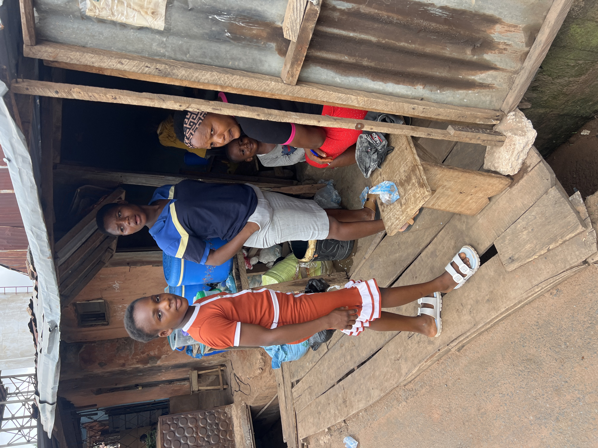 This image, taken in Obosi, Nigeria, captures four individuals amidst a rustic, makeshift structure, likely a dwelling or a market stall. The scene features a raised wooden platform and walls constructed from wooden planks and corrugated metal sheets.
In the background, positioned in a darker, recessed area under a low roof, an adult female with a patterned headscarf, black top, and red shorts smiles gently towards the camera. A young child with a light-colored top peeks out from behind her, also looking at the camera.
In the foreground, standing on the wooden platform, are two youths. On the left, a male youth or teenager wears a blue and yellow polo shirt and light-colored shorts, looking directly at the camera with a neutral expression. To his right, a female youth or teenager stands in an orange dress with white striped details and white sandals, also looking at the camera.
The immediate surroundings include more wooden planks, plastic bags, and blue plastic containers. The ground beyond the wooden platform appears to be a mix of dirt and possibly rough concrete. The lighting suggests it is daytime. There is no clearly discernible text visible on any objects or clothing.