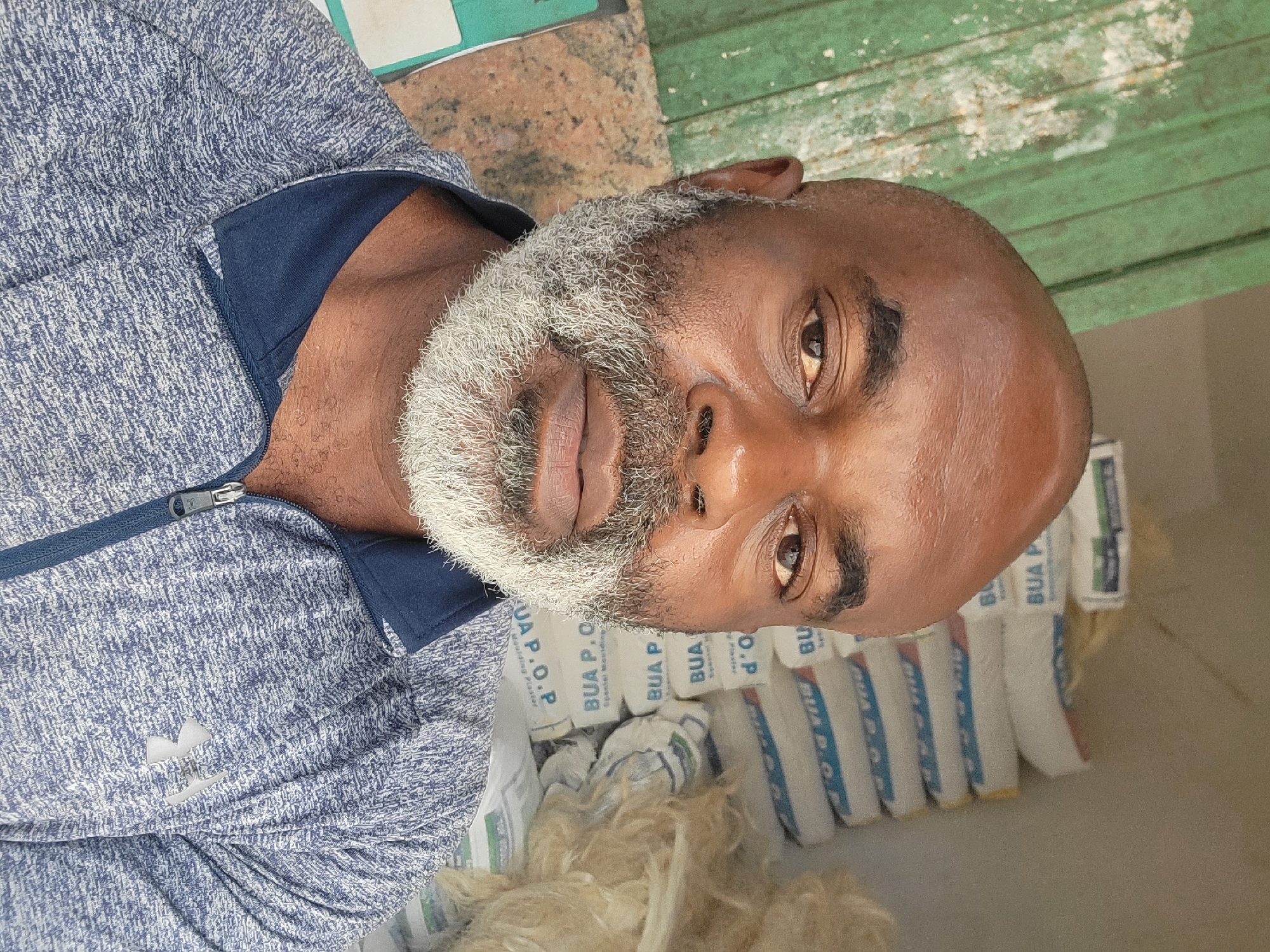 The image shows a man with a bald head and a gray beard wearing a blue and white jacket. He is positioned next to a stack of white bags labeled "BUA P.O.P." There is some light brown fibrous material in the foreground. The location in the city of Maiburiji, Nigeria is unknown. The location is unclear.
