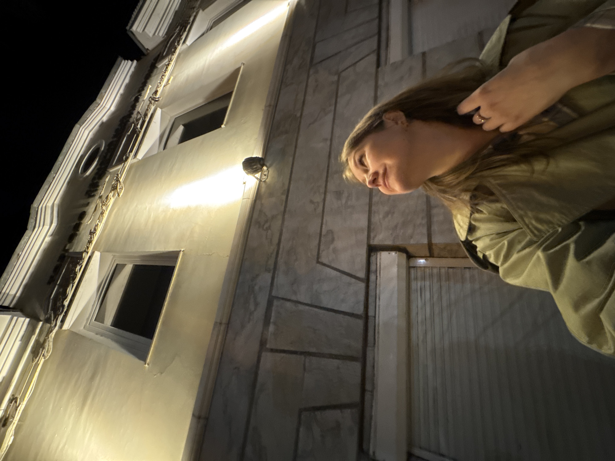 A low-angle shot captures a woman in a green jacket, positioned against a building facade illuminated by artificial light. The building's exterior features windows and marble-like paneling, creating a stark contrast with the black sky in the background. A cylindrical street light fixture is mounted on the building's wall. The location is in the city of Perm, Russia.