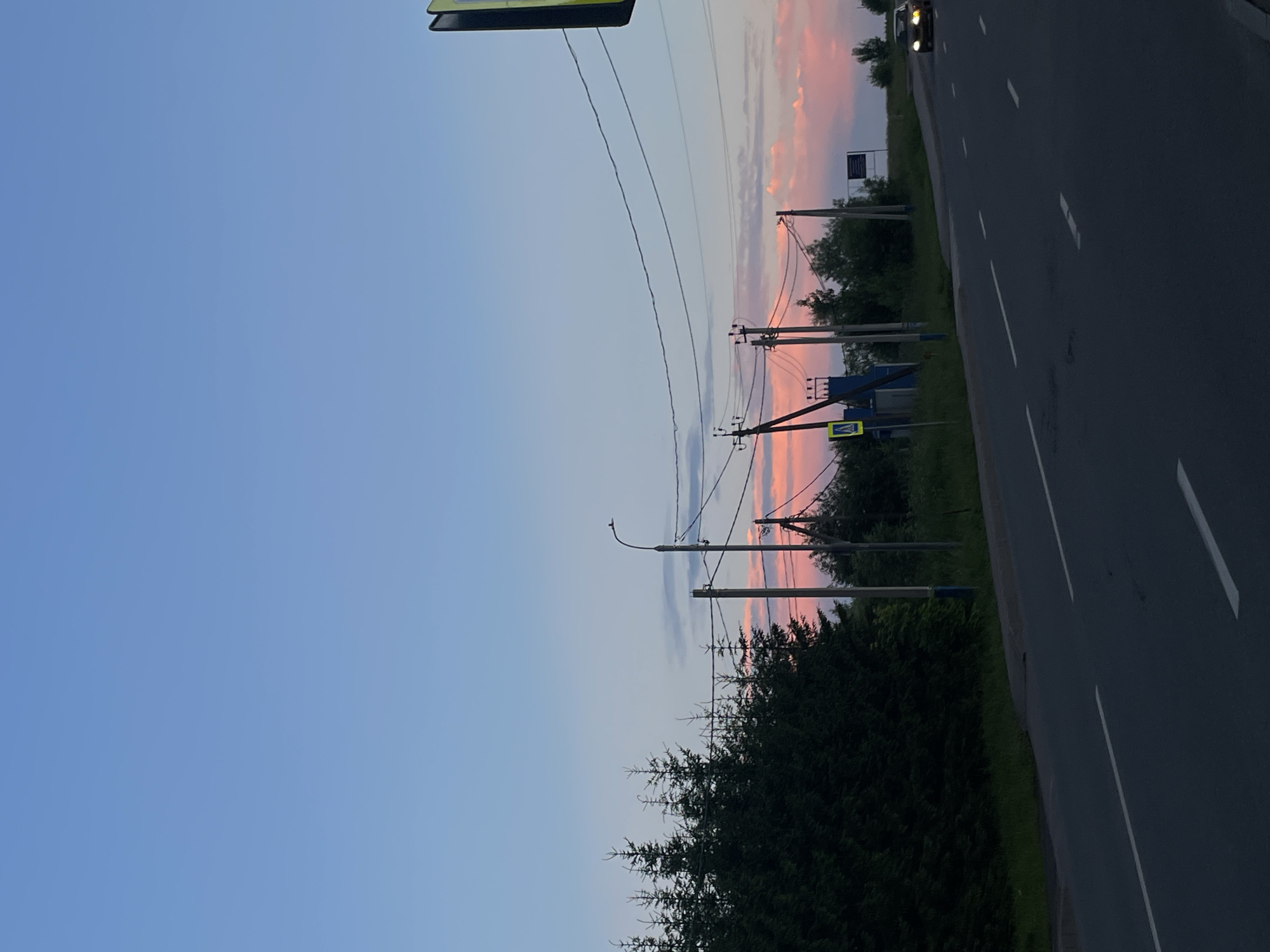 The image captures a road scene at dusk in the city of Zverevo, Russia. The sky displays a gradient of soft blue to pink hues, indicative of a sunset. Along the left side of the road, a dense line of dark green coniferous trees stands tall. A paved road, marked with white dashed lines, extends into the distance, curving slightly to the right. Several utility poles with visible wires are positioned along the roadside, one of which supports a street lamp. A pedestrian crossing sign with a yellow triangular warning symbol is visible near the poles. In the distance, on the right side of the road, a single car with illuminated headlights is driving away from the viewer. The overall environment appears to be semi-rural or on the outskirts of a town, with elements of infrastructure like power lines and road markings juxtaposed against the natural backdrop of trees and a colorful sky.