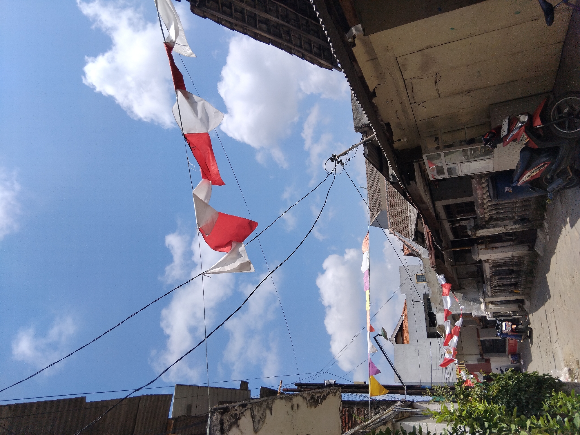 The image depicts a street scene in the city of Kepuh Kiriman, Indonesia, on a sunny day with a blue sky and scattered clouds. Red and white Indonesian flags are strung across the street, adorning the buildings and overhead wires. The architecture suggests a residential or mixed-use area, with buildings featuring corrugated metal roofs and some showing signs of wear.
On the left side of the street, a building with weathered concrete walls and exposed rebar is visible, with green foliage growing at its base. Further down the street, a narrow pathway leads past a row of buildings, some of which have fences or walls.
Several motorcycles are parked on the street. One red motorcycle is prominently parked in the foreground on the right side of the image, next to a beige-colored building with a large, sliding door. Another motorcycle, partially visible, is parked further down the street in the middle ground.
The scene is calm and devoid of human activity, capturing a snapshot of everyday life in this Indonesian locality, possibly during a festive occasion given the abundance of national flags.