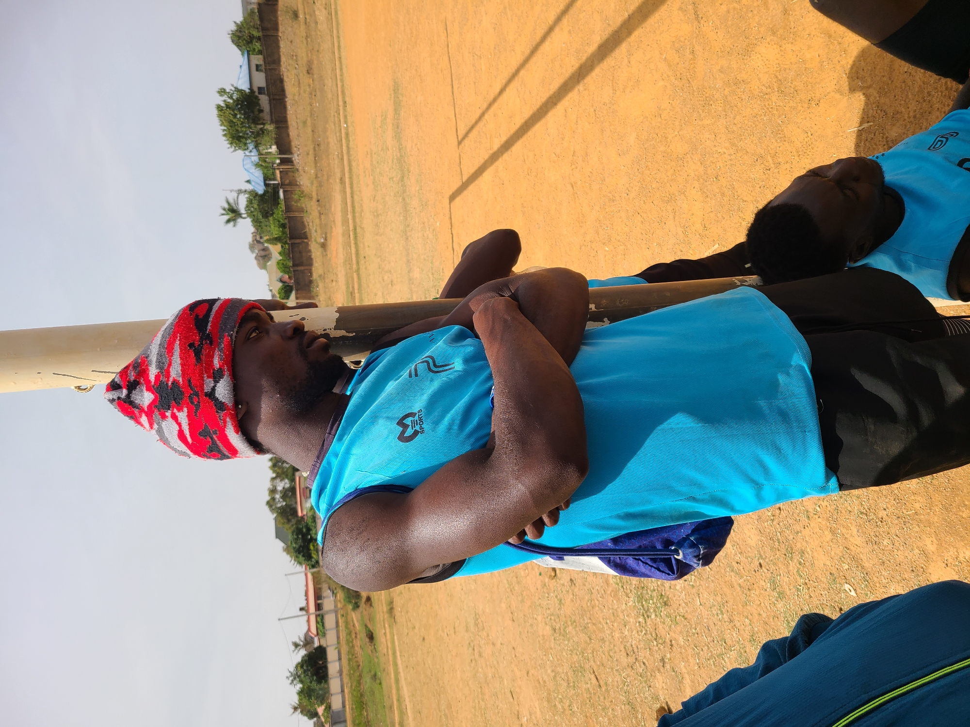 The image captures an outdoor scene featuring two men resting on what appears to be a dusty sports field or open ground. The foreground subject, prominently displayed, is a man with dark skin, a beard, and wearing a red, black, and grey knit beanie. He is dressed in a bright blue short-sleeved athletic shirt, which has "M SPORTS" printed on the right chest area and another abstract logo on the left. He also wears dark shorts or trousers. His arms are crossed over his chest, and his skin exhibits a sheen, suggesting he may be sweaty. He leans against or lies beside a horizontal metal pole, looking slightly upwards or to the side.
Another man is partially visible in the mid-ground, also resting on the ground, wearing a similar blue athletic shirt and dark shorts. His face is mostly obscured. The field itself is a light brown-orange, dry and dusty, with subtle markings that could indicate a playing area. In the background, under a clear, bright sky, are several buildings with blue roofs, scattered green trees including palm trees, and some rock formations, characteristic of a natural or semi-rural environment.
The strong sunlight and clear shadows cast by the pole indicate it is daytime, likely mid-day or early afternoon. The men's relaxed postures and athletic attire suggest they are either taking a break from physical activity or simply lounging in the warm, sunny weather. A blue bag is partially visible near the foreground man, and part of another person's leg in dark blue trousers with a green stripe can be seen on the far right. The scene is consistent with an outdoor sports or leisure activity in Barikin Jirga Summa, Nigeria.