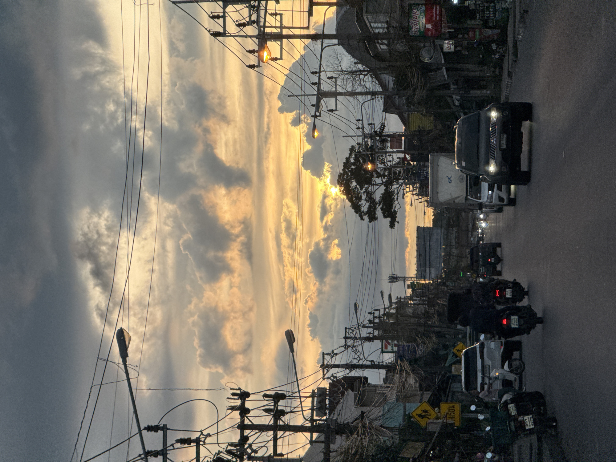 This image depicts a street scene at dusk in Baan Mae Nam, Thailand. The sky is filled with dramatic clouds tinged with the warm, golden hues of the setting sun, creating a picturesque backdrop. Power lines and utility poles crisscross the frame, a common sight in the region, adding a sense of urban infrastructure to the natural beauty.
The street is busy with traffic. Several motorcycles are visible, some with riders, suggesting a typical evening commute or activity. A dark-colored SUV is in the foreground, its headlights illuminated. A white truck is also present, along with other vehicles further down the road. The presence of these vehicles indicates that this is a public roadway, likely a main thoroughfare within the town.
Signs with Thai script are visible on the buildings lining the street, confirming the location in Thailand. One yellow sign with black symbols is a traffic sign indicating a school crossing. The overall mood is one of a calm, yet active, end to the day, with the dramatic sky adding an element of awe. The low light suggests it is either sunrise or sunset, but the activity of vehicles and the golden sky strongly indicate sunset.