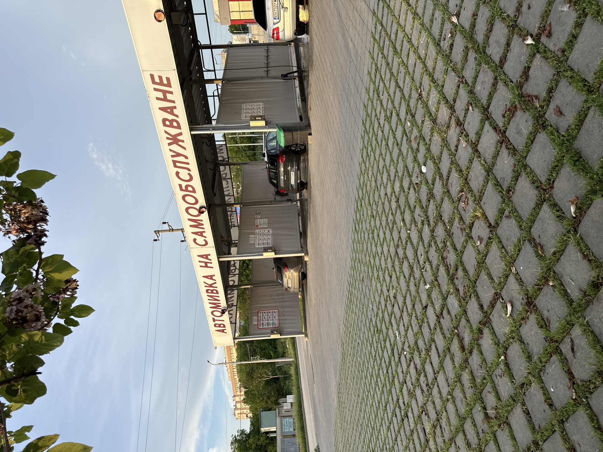 The image shows a self-service car wash ("Автомивка на самообслужване") in Pleven, Bulgaria. The car wash consists of multiple bays with gray walls and a metal structure above. Cars are parked in several of the bays. The ground is paved with gray concrete blocks with grass growing in the spaces between the blocks. The sky is visible with some clouds. Trees are present in the background and to the left of the frame. Power lines run above the car wash. A flower tree sits to the left of the image.