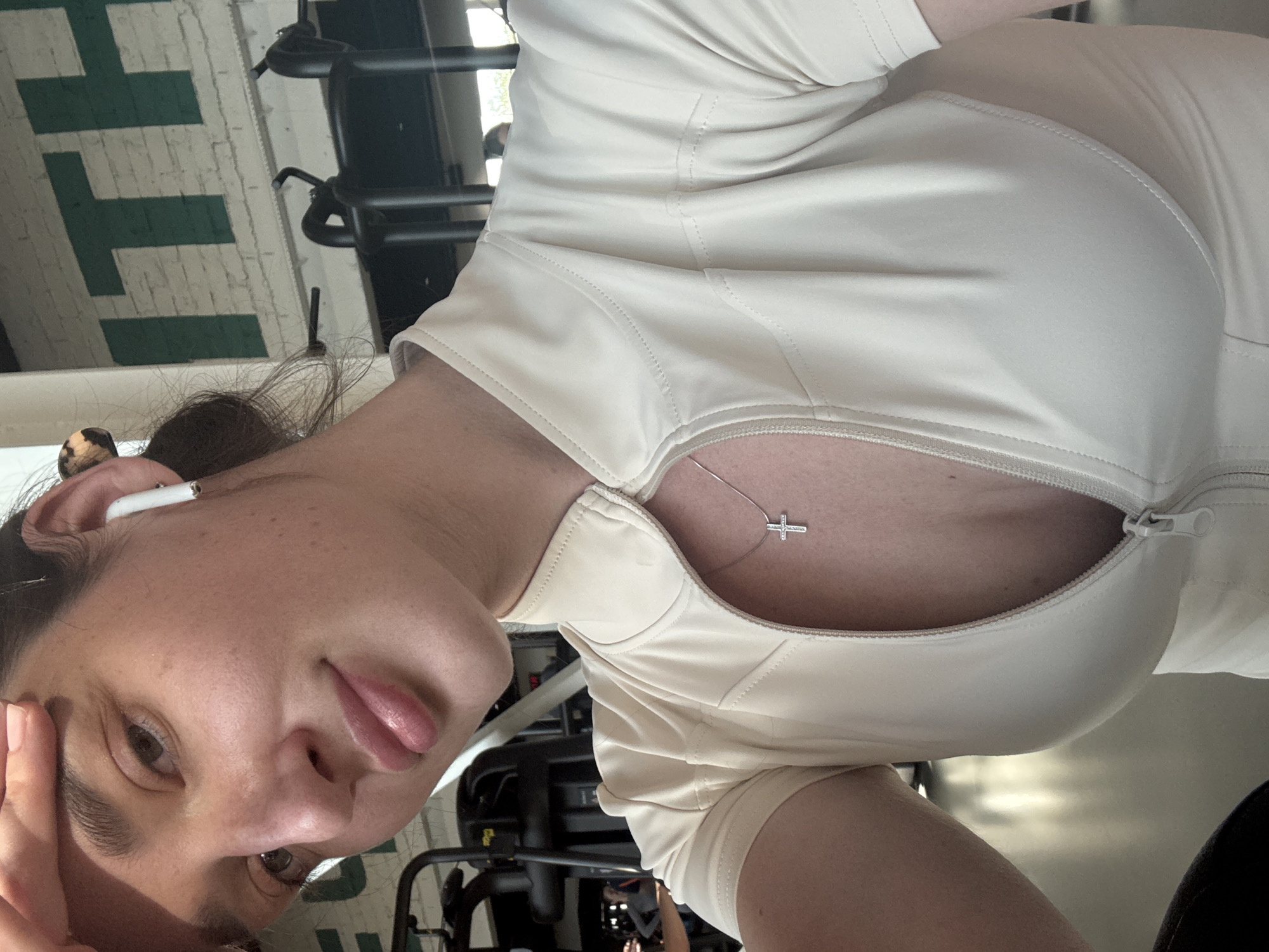 A selfie shows a woman wearing a beige zippered top and a delicate cross necklace, positioned in front of gym equipment. She has dark hair pulled back, an earbud in her ear, and is resting her head on her hand. The backdrop includes indistinct gym machines and a wall marked with green and white architectural elements.