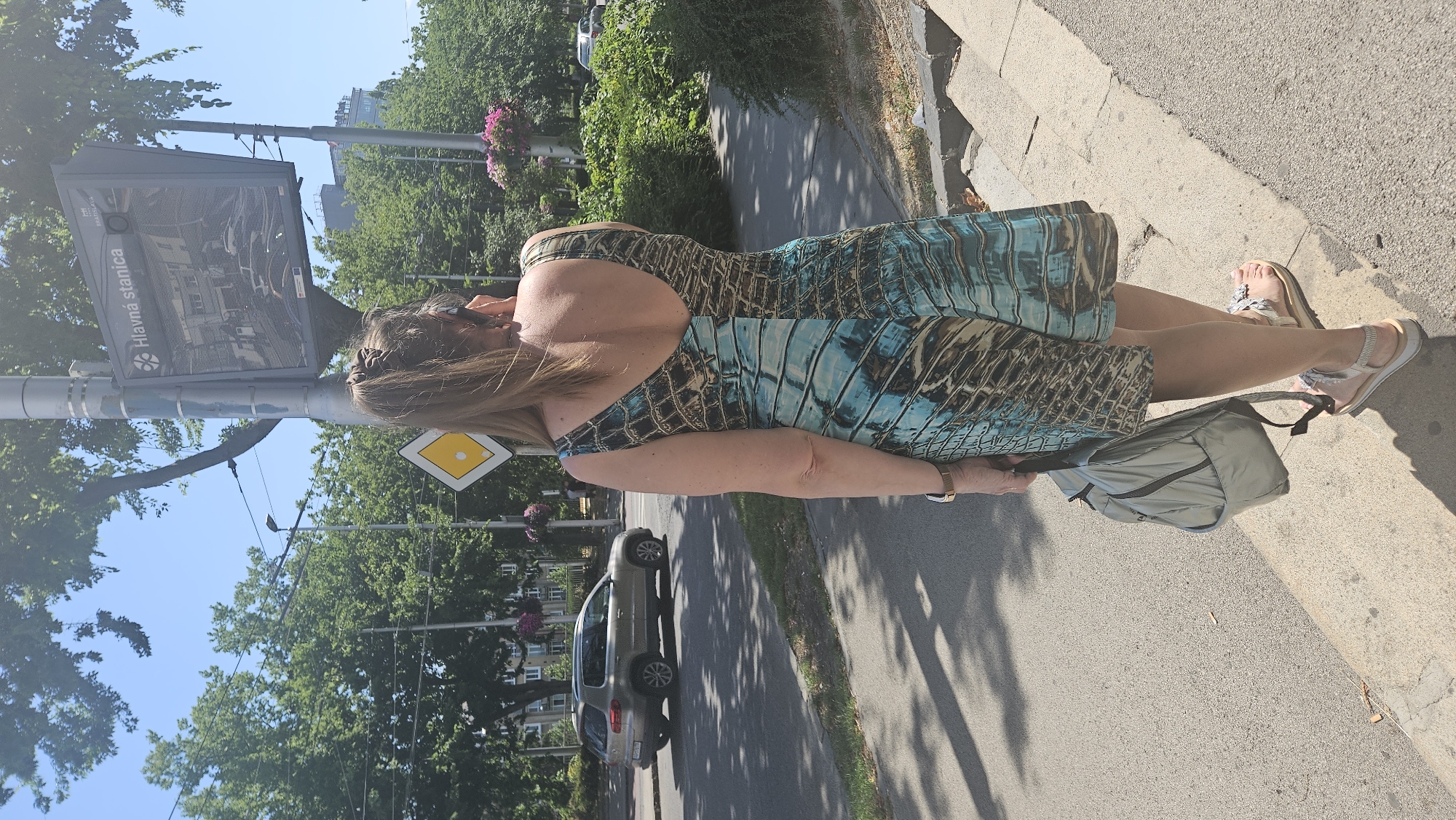 The image shows a woman from behind, wearing a patterned dress and sandals, holding a backpack. She is standing on a sidewalk in Bratislava, Slovakia. To her left, a car is driving on a street. In the background, there are trees and buildings, and a digital display board with the text "Hlavná stanica" which translates to "Main Station" in Slovak. The digital board also displays a map and route information for public transport. The weather is sunny and clear.