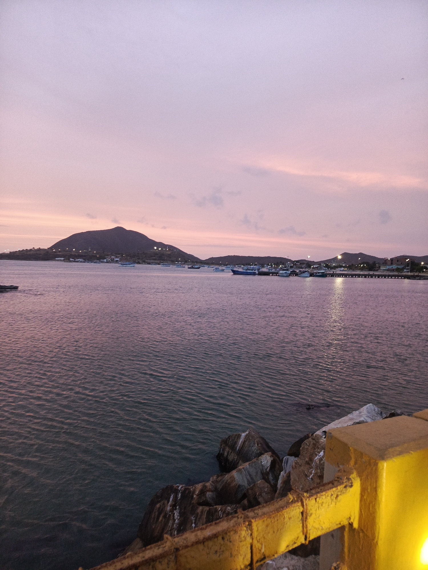 The image depicts a view of the bay in Juan Griego, Venezuela, taken at dusk. The sky is a blend of pink and purple hues. In the distance, there's a prominent hill or small mountain. The bay itself is calm, reflecting the colors of the sky. Several boats are anchored in the bay near the shore. The edge of the pier or seawall is in the foreground, with large rocks and a yellow metal railing visible. The railing is partially illuminated, likely by a nearby light source.