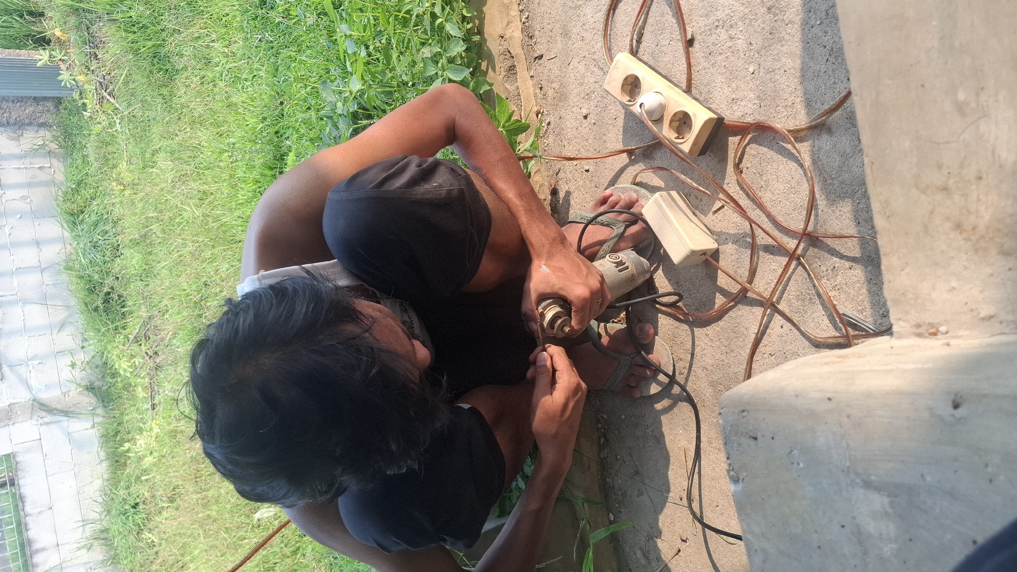 A man is crouched on a concrete surface outdoors, working with a small, handheld electric rotary tool. He is wearing black shorts and a white tank top, with green flip-flops on his feet. His hair is dark and appears to be slicked back. In front of him are several electrical components, including a power strip with three outlets, a small beige electrical box, and various coiled electrical cords. The background consists of green grass, a concrete wall, and some indeterminate greenery. The lighting suggests it is daytime, with sunlight casting shadows. The scene appears to be in a residential outdoor area, possibly a backyard or alleyway. The specific location in Rahong, Indonesia, is not discernible from the visual evidence alone, but the overall environment is consistent with a tropical or subtropical climate.