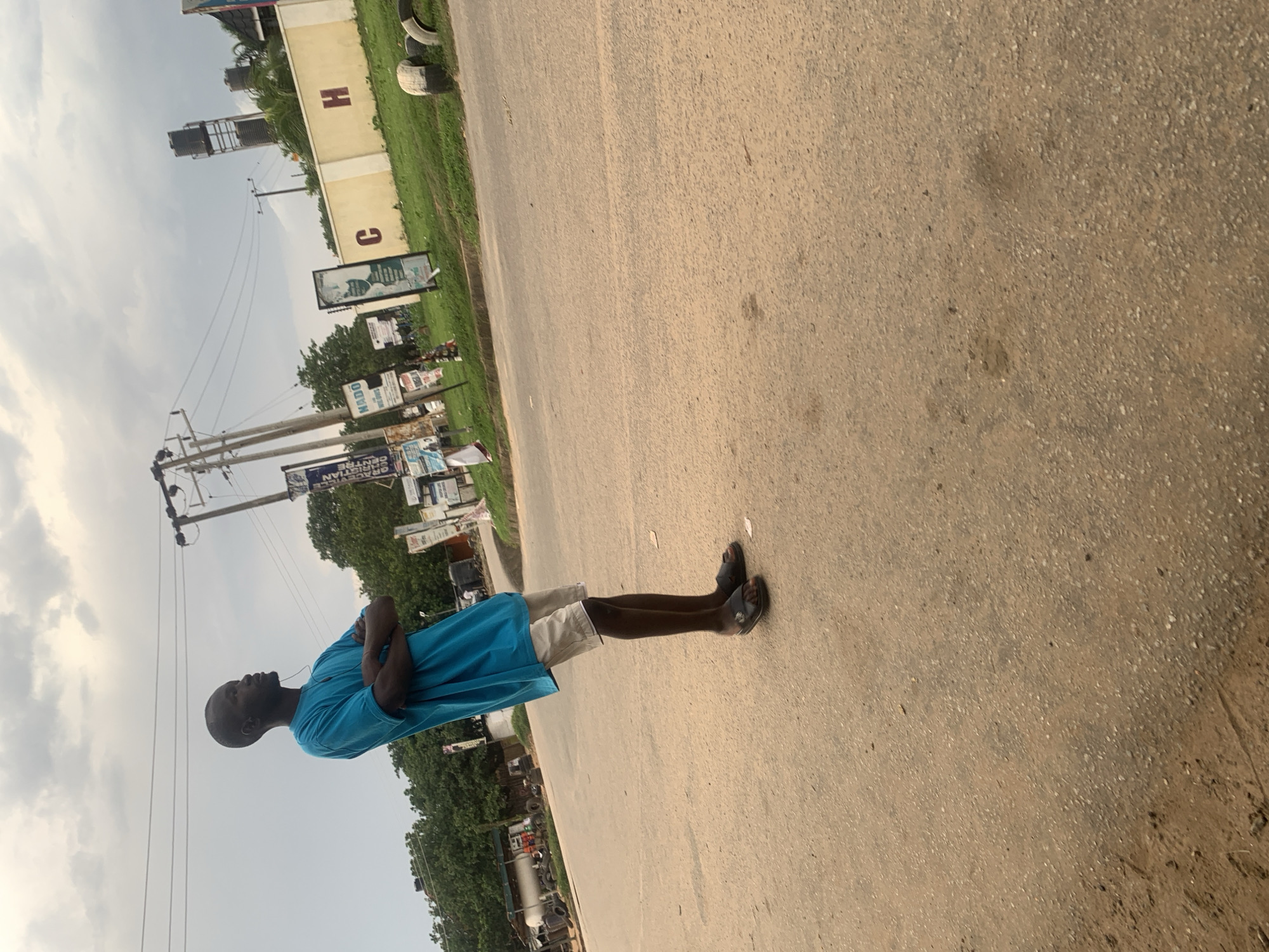 A young man stands with his arms crossed, facing away from the camera and looking to his right, near a road in Abuja, Nigeria. He is wearing a bright blue t-shirt and light-colored shorts, with black sandals. The ground is paved with rough asphalt. In the background, there are trees, utility poles with power lines, several billboards, and a yellow wall with the letters "C" and "H" visible. There are also water tanks visible on a tall structure in the distance. The sky is overcast.