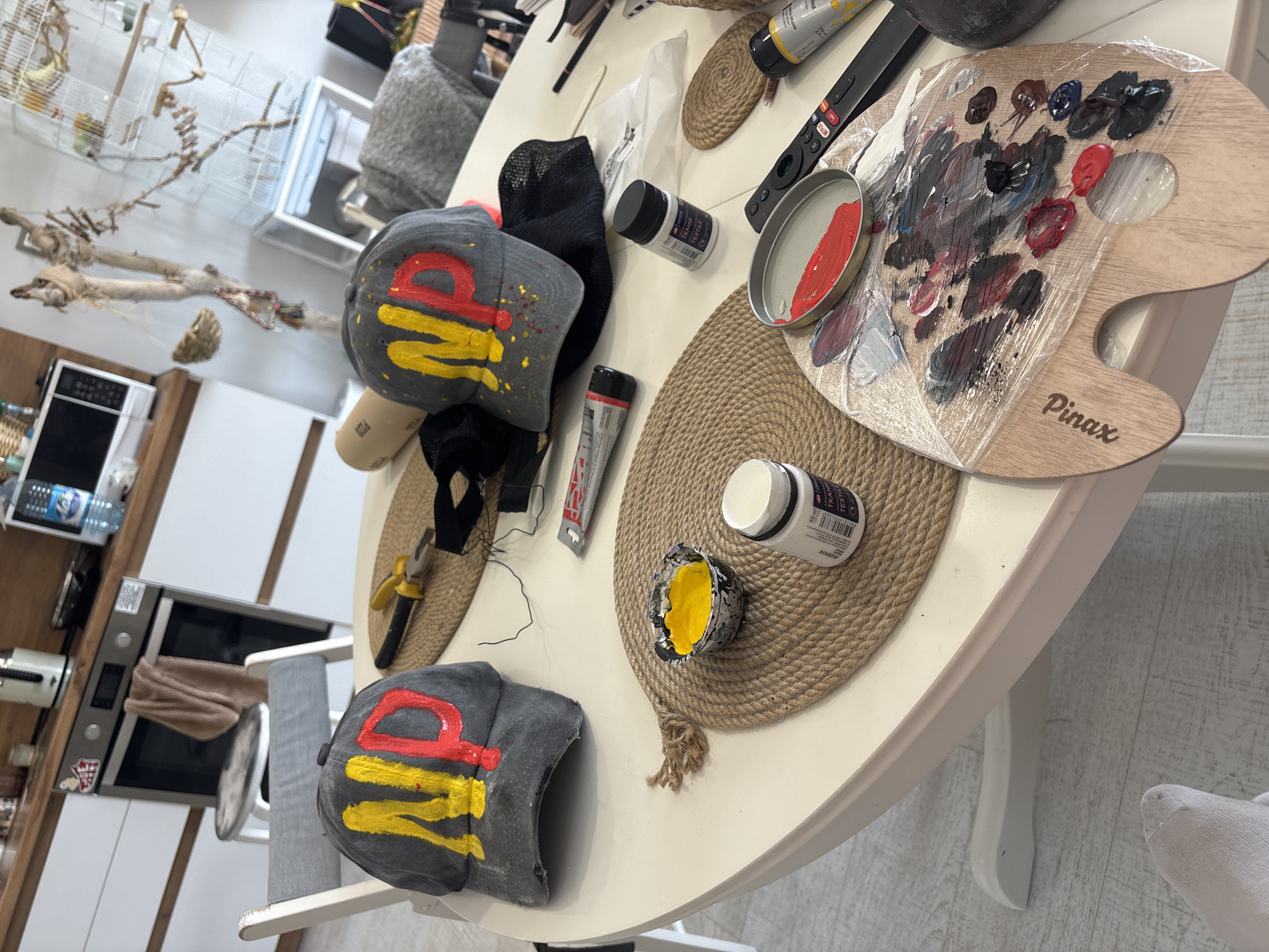 The image shows a table in what appears to be a domestic setting, possibly in Krasnoyarsk, Russia, based on Cyrillic text visible on some of the objects. The table is round and white, with several crafting items arranged on it. There are two gray baseball caps with stylized red and yellow paint designs. Several containers of paint are visible, including tubes and jars. A wooden artist's palette covered in what appears to be plastic wrap, and smeared with various colors of paint, is also on the table. Other items include a pair of pliers, some black fabric, and circular woven placemats. The background includes kitchen appliances and a birdcage.