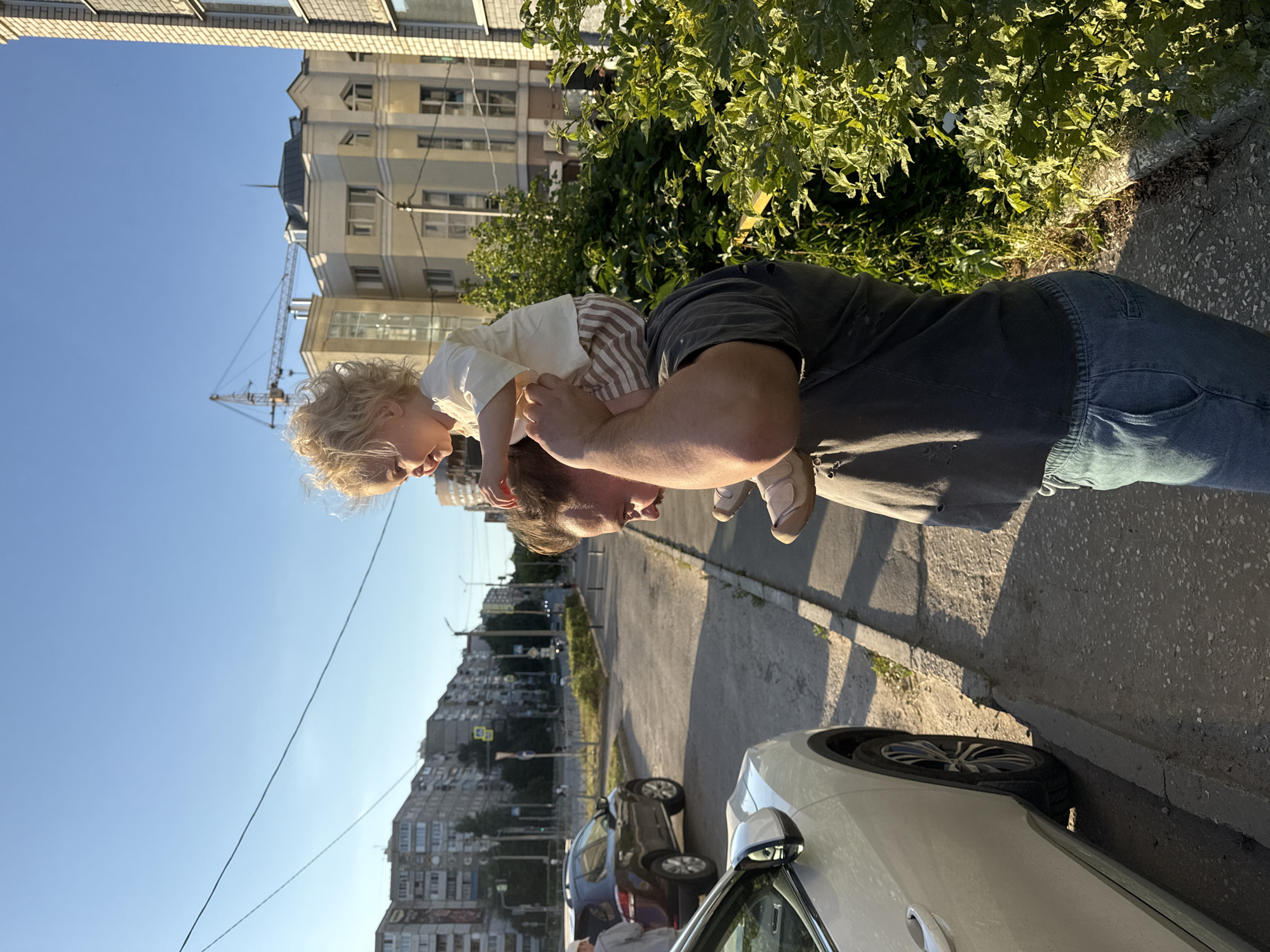 In this photograph, a man carries a young child on his shoulders. The child, with blonde curly hair, wears a light-colored shirt and striped shorts. They are both looking towards the left side of the frame. The man is wearing a dark t-shirt and blue jeans. The scene appears to be outdoors, with a sidewalk and street visible. Several cars are parked along the street, and a building with multiple windows is in the background. Lush green foliage is also present. The lighting suggests it is daytime, likely late afternoon, given the long shadows. The sky is clear and blue.
Visible text: There is no visible text in this image.