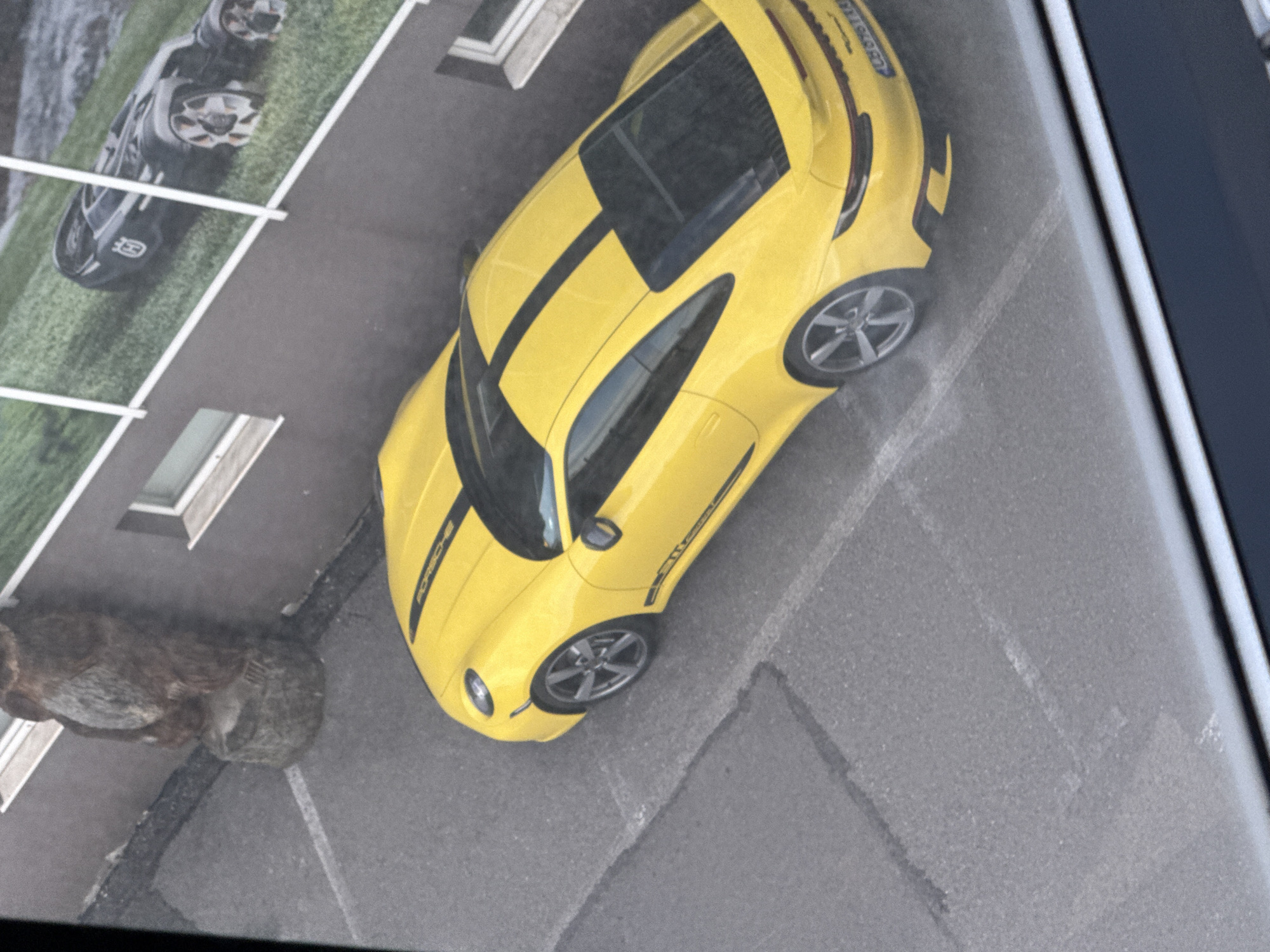 The image depicts a print of a yellow Porsche with black stripes parked on a paved surface, possibly a street. The scene includes parts of buildings with windows and another car depicted in the background. A statue or large object resembling a weathered animal is also visible near the yellow Porsche. The overall angle of the image suggests it's a photograph of a print or poster hanging somewhere. There's no direct evidence in the image to suggest it's specifically in Sofia, Bulgaria; it could be anywhere with similar architectural features.