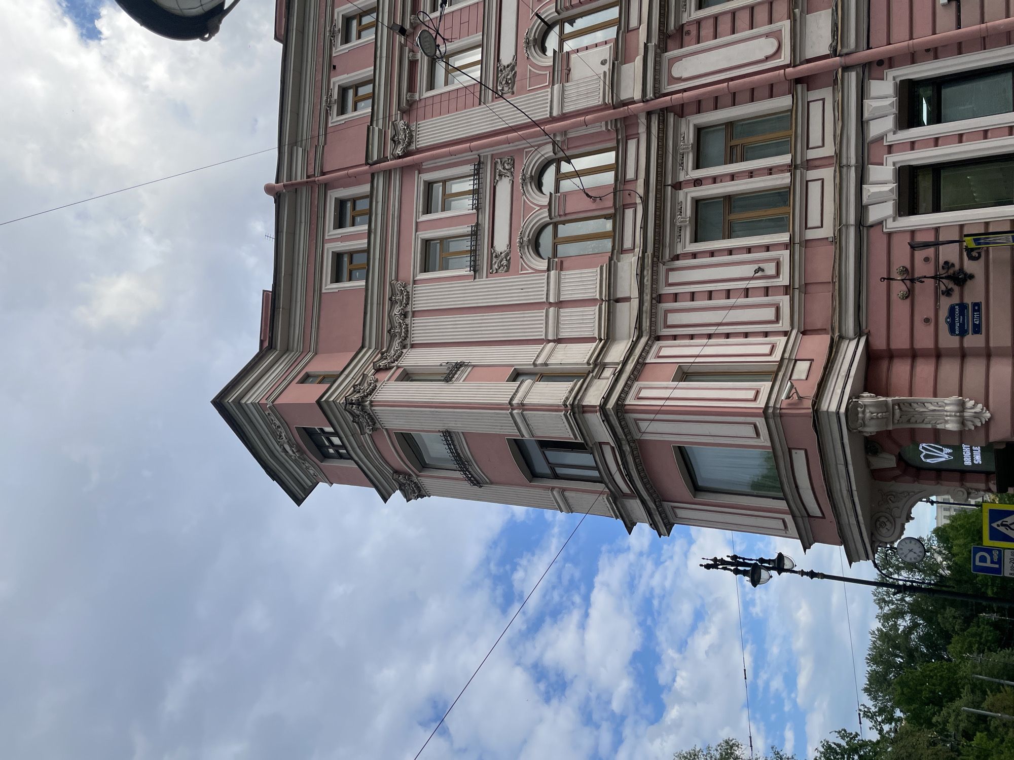 This image presents an upward, angled view of a large, ornate building under a partly cloudy sky. The building's facade is a prominent pink, richly decorated with white or light-colored architectural details, including pilasters, decorative friezes, and moldings. It features a variety of windows, both rectangular and arched, some with rounded frames and small dark grates. Rain gutters and electrical wires are visible running along the building's exterior.
The sky above is filled with scattered white and grey clouds, indicating a daytime setting with moderate cloud cover. Numerous overhead wires traverse the frame, connected to the building and nearby utility poles.
In the lower right section of the image, elements of urban street furniture are visible. These include a tall, black lamppost with multiple ornate light fixtures, a segment of a round clock face, and the green foliage of trees in the background.
Several signs with visible text offer specific location cues. A vertical black sign with a white logo reads "BRIGHT SMILE," indicating a business. Below it, a blue street sign in Cyrillic displays "ФУРШТАДСКАЯ" (Furshtadskaya), identifying the street, along with the building numbers "47/11." Another blue sign featuring a white "P" denotes parking, with "8.00-20.00" specifying the operational hours.
The scene is static, capturing the architectural beauty and urban environment of what is identified by the Cyrillic script and context as Saint Petersburg, Russia.