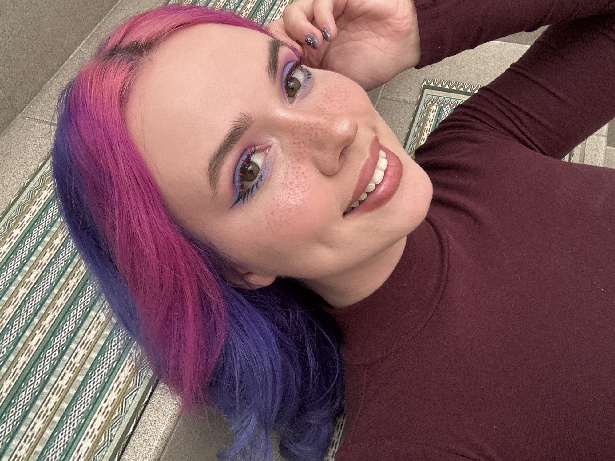 A close-up, high-angle selfie captures a young woman lying down and smiling broadly, engaging directly with the camera. She has vibrant, split-dyed hair, with bright pink on the side closer to the camera and purple on the other, transitioning from the mid-lengths. Dark roots are visible at her hairline on the pink side. Her light complexion is adorned with scattered freckles across her nose and cheeks. She has expressive brown eyes, which are accentuated by purple eyeshadow and distinctive black eyeliner featuring a spiky, graphic design along the lower lash line. Her lips are painted with a light pink or nude lipstick, revealing white teeth as she smiles. She wears a high-necked, long-sleeved top in a deep maroon or burgundy color. Her left hand, with nails painted in a dark, glittery polish, rests near her head. The background consists of a geometric patterned rug or mat featuring green, beige, white, and black hues, partially visible on a light-colored, possibly tiled or concrete floor. The overall impression is one of cheerfulness and self-expression. The scene is set in the City of Zagreb, Croatia. No visible text is present.