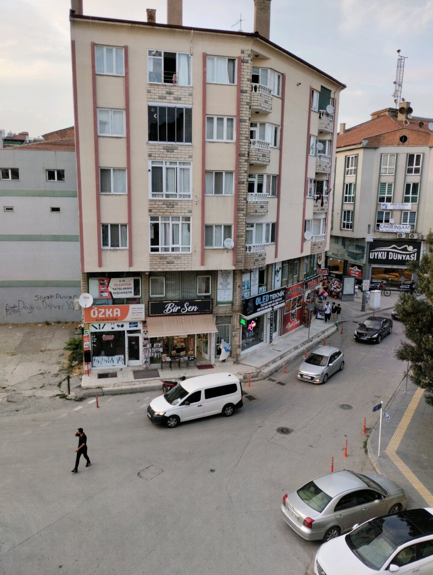 In Çorum, Turkey, an urban street scene unfolds with buildings and traffic. A multi-story beige building with red trim dominates the center, its facade featuring numerous windows and balconies. The ground floor of this building houses several businesses, including "ÖZKA ELEKTRONIK" and "Bir Sen." Adjacent to it, another building with a tiled roof and multiple windows is visible.
The street below is paved and shows signs of wear, with manhole covers and painted lines. Several vehicles are present: a white van is parked on the street, a silver sedan is in motion, and a black sedan is also on the road. A lone pedestrian, dressed in dark clothing, walks purposefully across the street. The environment suggests a moderately busy urban area, with utility poles and antennas visible on rooftops. The overall lighting indicates it is daytime, possibly late afternoon or early evening.