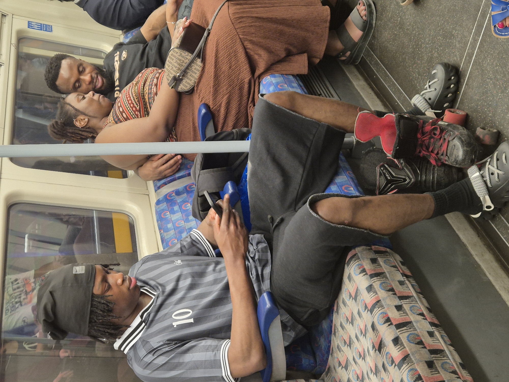 The image captures a scene inside what appears to be a train carriage, likely part of the London Underground or a similar urban railway system in London, United Kingdom.
Several people are present. A man wearing a grey-striped shirt with the number "10" on it sits facing the camera, looking at a device in his hands. The seat beside him is patterned.
Next to him, a couple occupies two seats. The man is wearing a black t-shirt and embraces the woman, who is wearing a patterned top. Another person is partially visible to the left, while a woman is seen on the upper right wearing a brown dress. Next to her is a pair of hockey skates.
The interior of the carriage is visible with overhead structures, windows, and a horizontal support bar. The seating fabric has a distinct pattern, indicative of London's public transportation. The lighting in the carriage is somewhat muted.