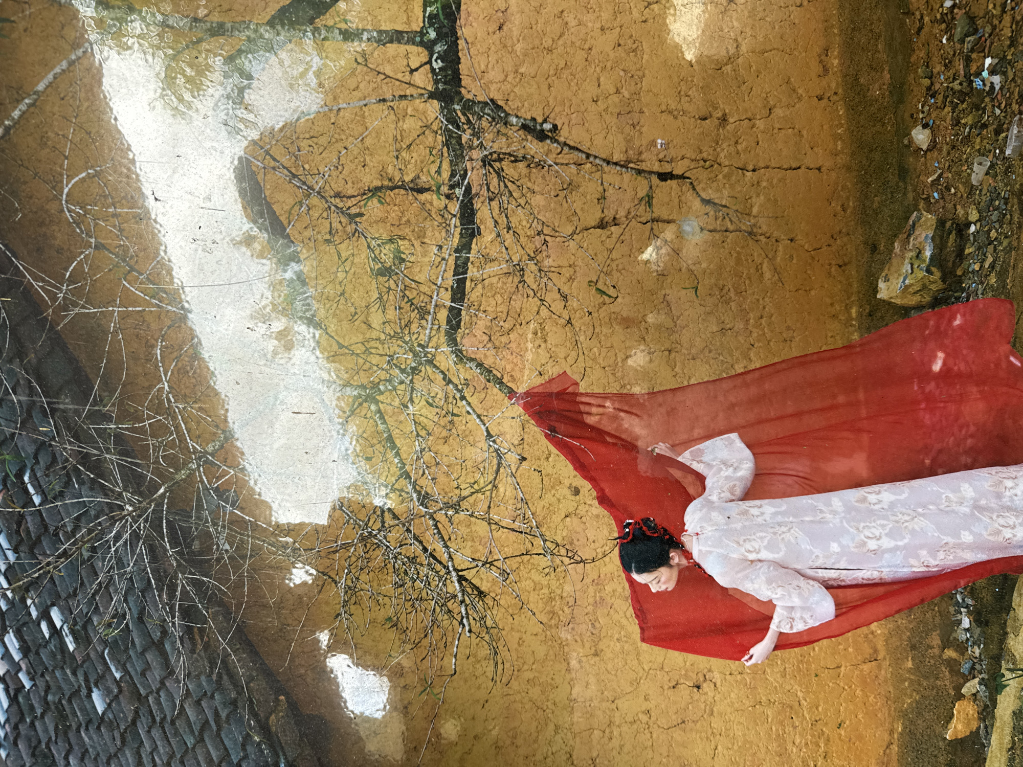 The image depicts a woman in traditional Vietnamese clothing, lying on a red sheet in what appears to be an outdoor setting in the Lũng Cú Commune, Vietnam. The background features a building with tiled roof, a bare tree, and what seems to be a dirt or clay-like surface. There is visual evidence of water or a wet surface, reflecting some of the scene. Small rocks and debris are scattered near the edge of the red sheet. The woman is positioned diagonally in the frame, with her head towards the lower left and feet extending towards the upper right.